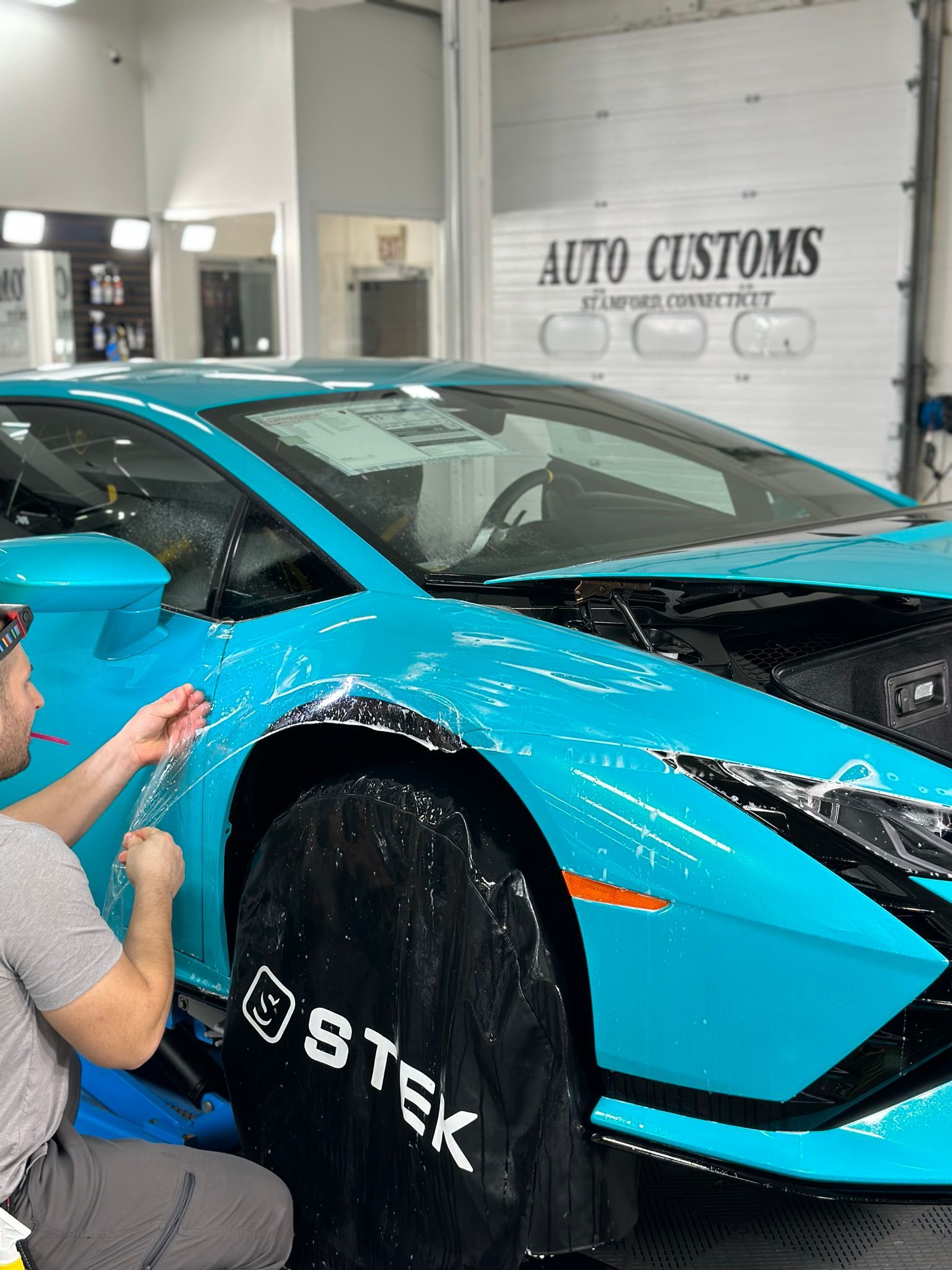 Man applying protective film to a bright blue Lamborghini in a garage.