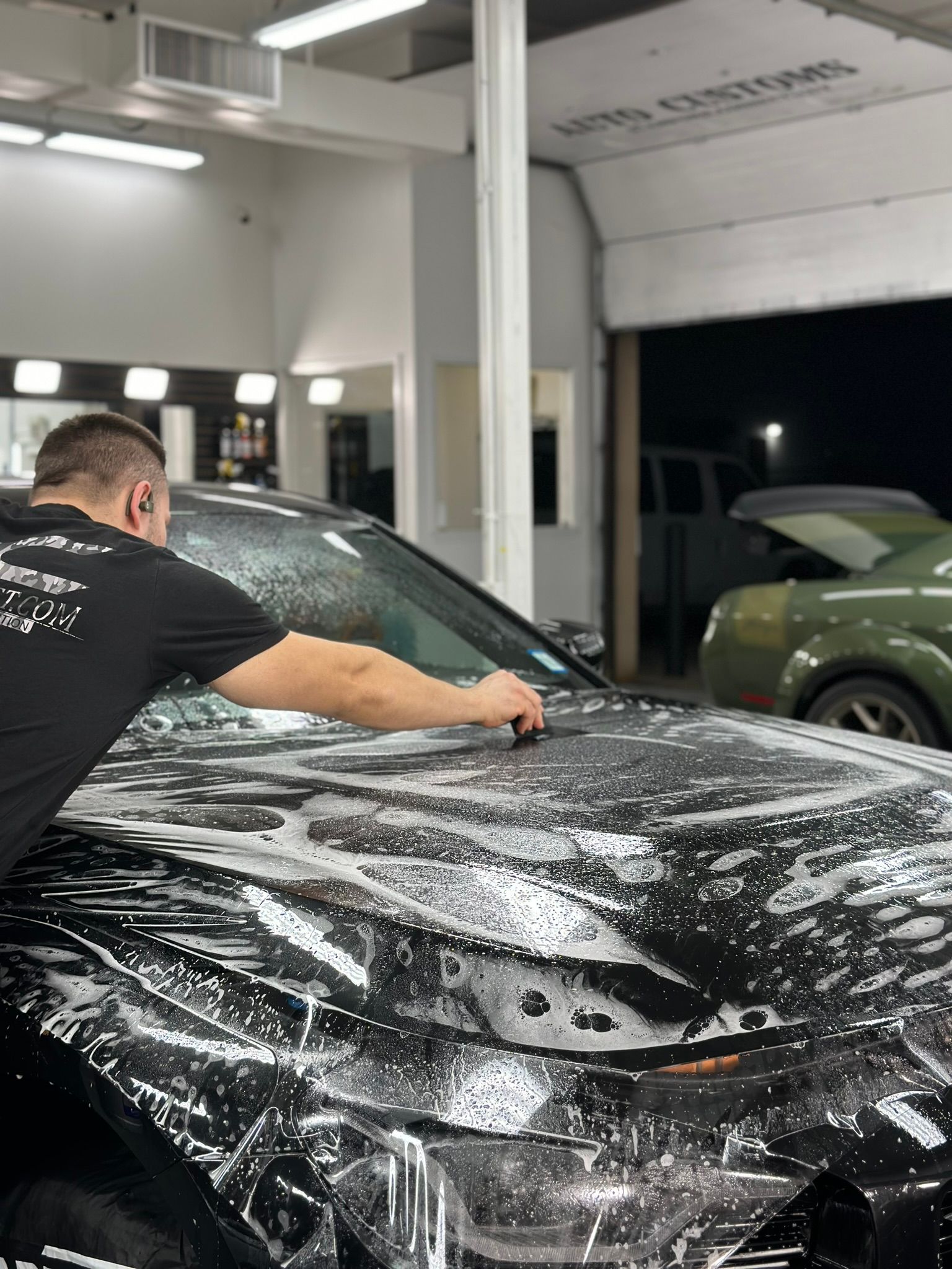 Man applying soapy film to black car hood in garage. Another green car in the background.