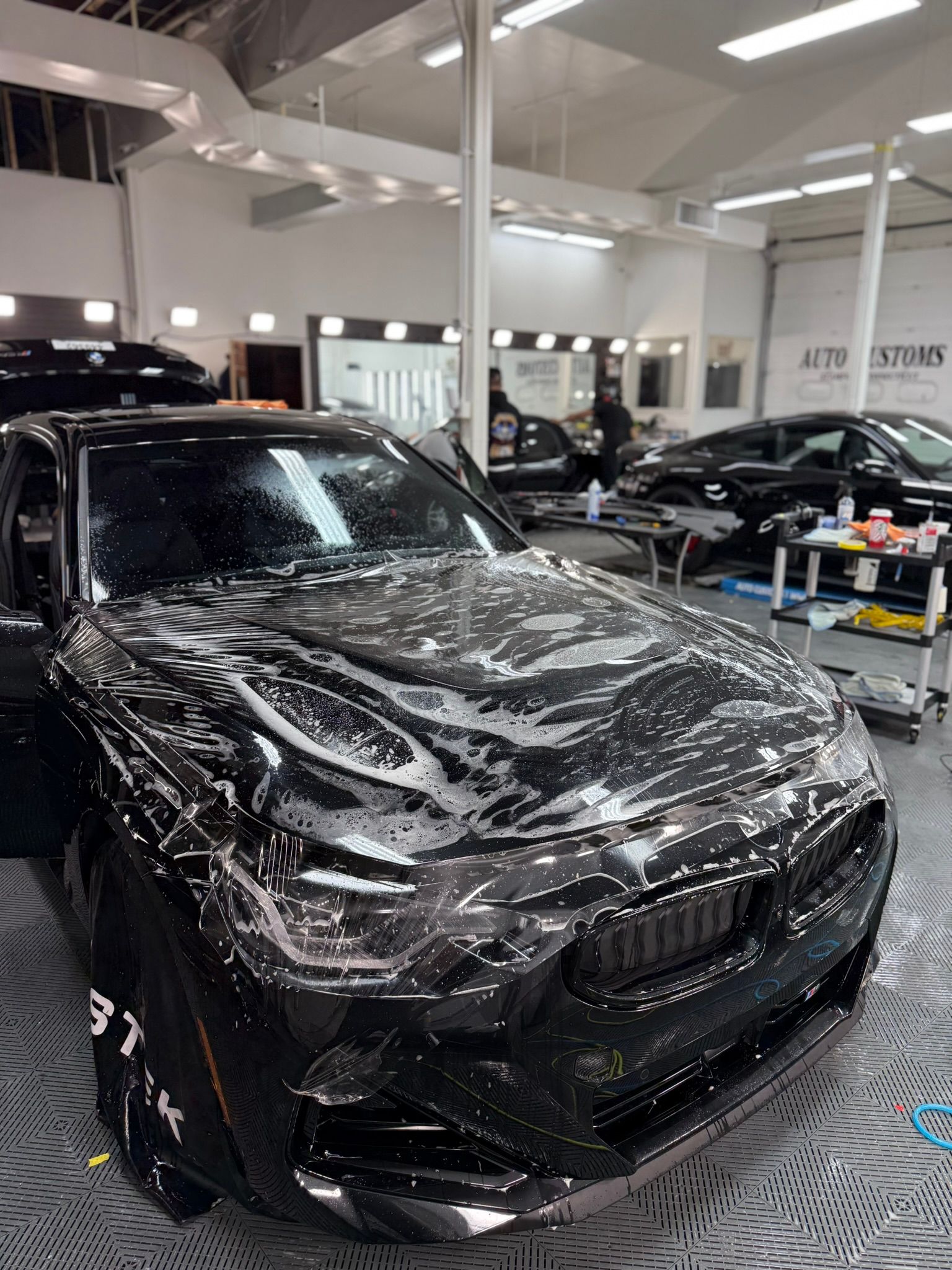 Black car covered in soapy water, inside auto shop. Person applying film.