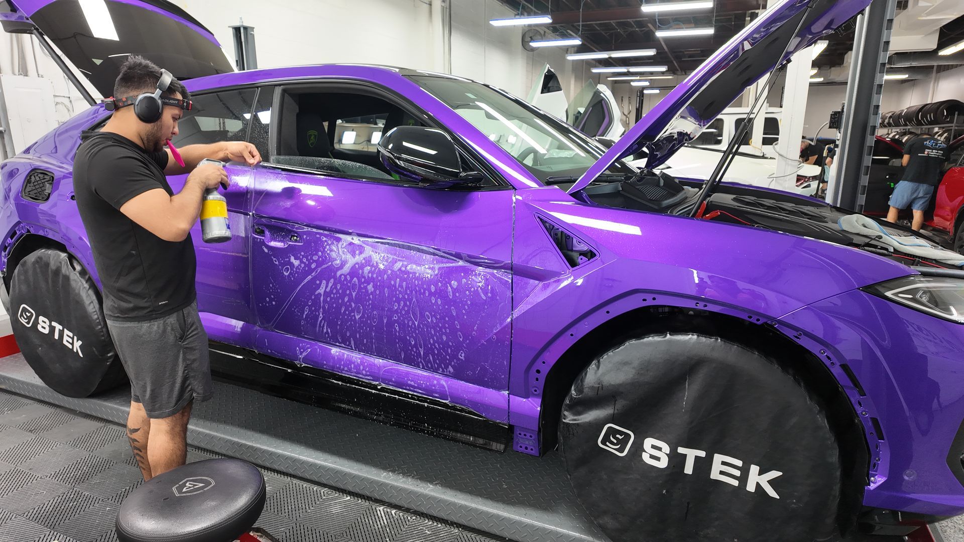 Man spraying purple Lamborghini in shop; hood and door open; black wheel covers.