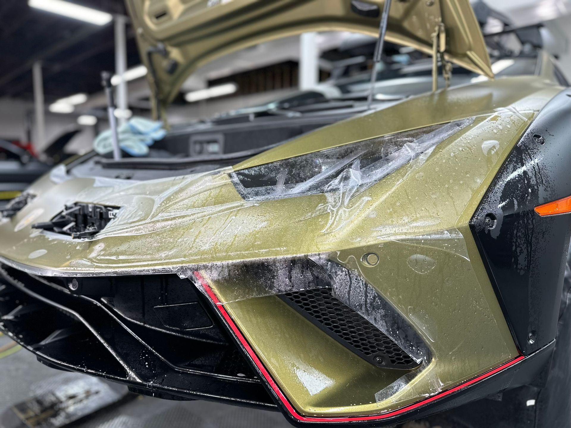 A Lamborghini's front end during paint protection film application. Gold-green paint, black accents, and red trim.