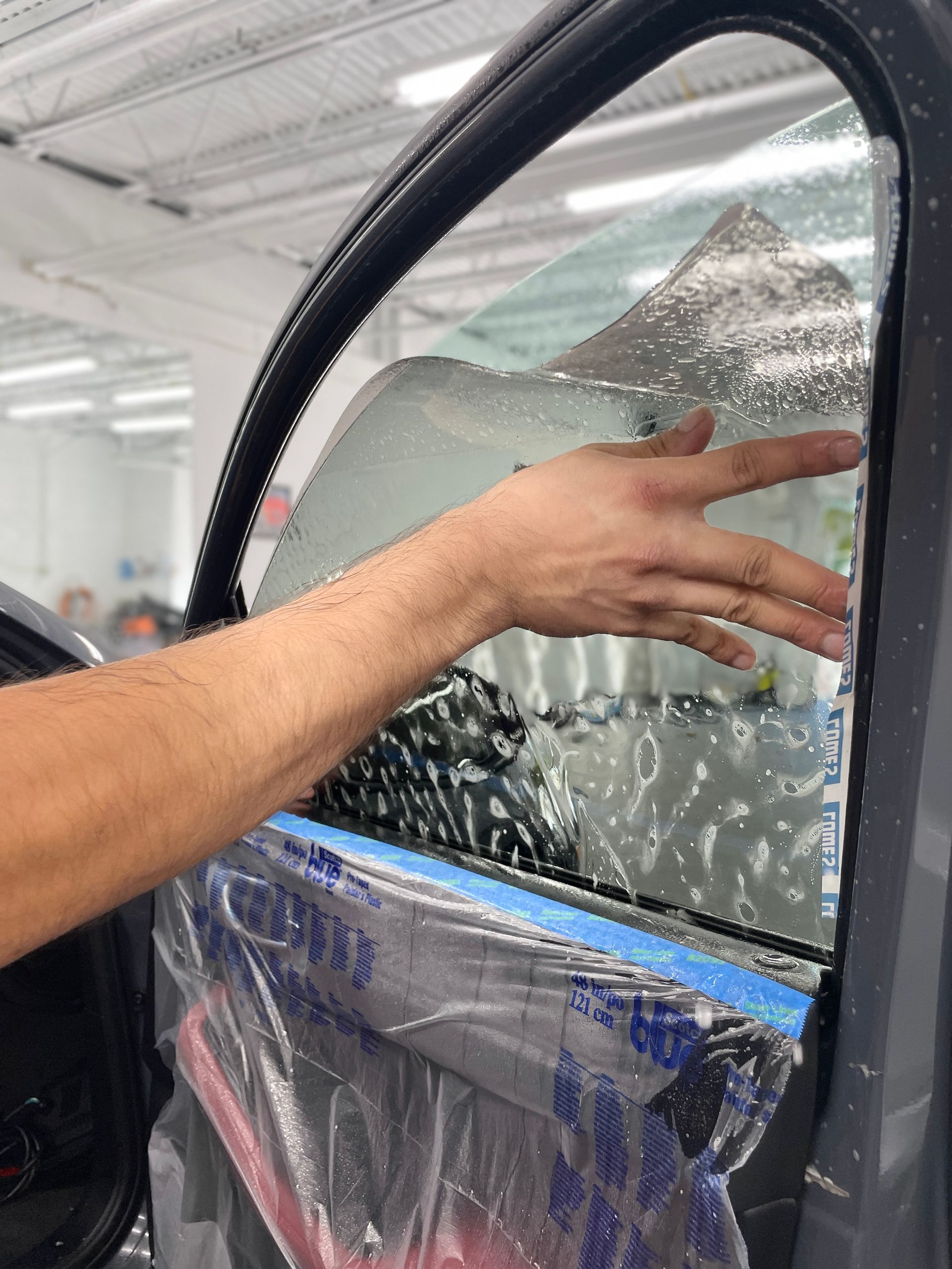 Person applying tinted film to a car window in a shop; hand smoothing the film.