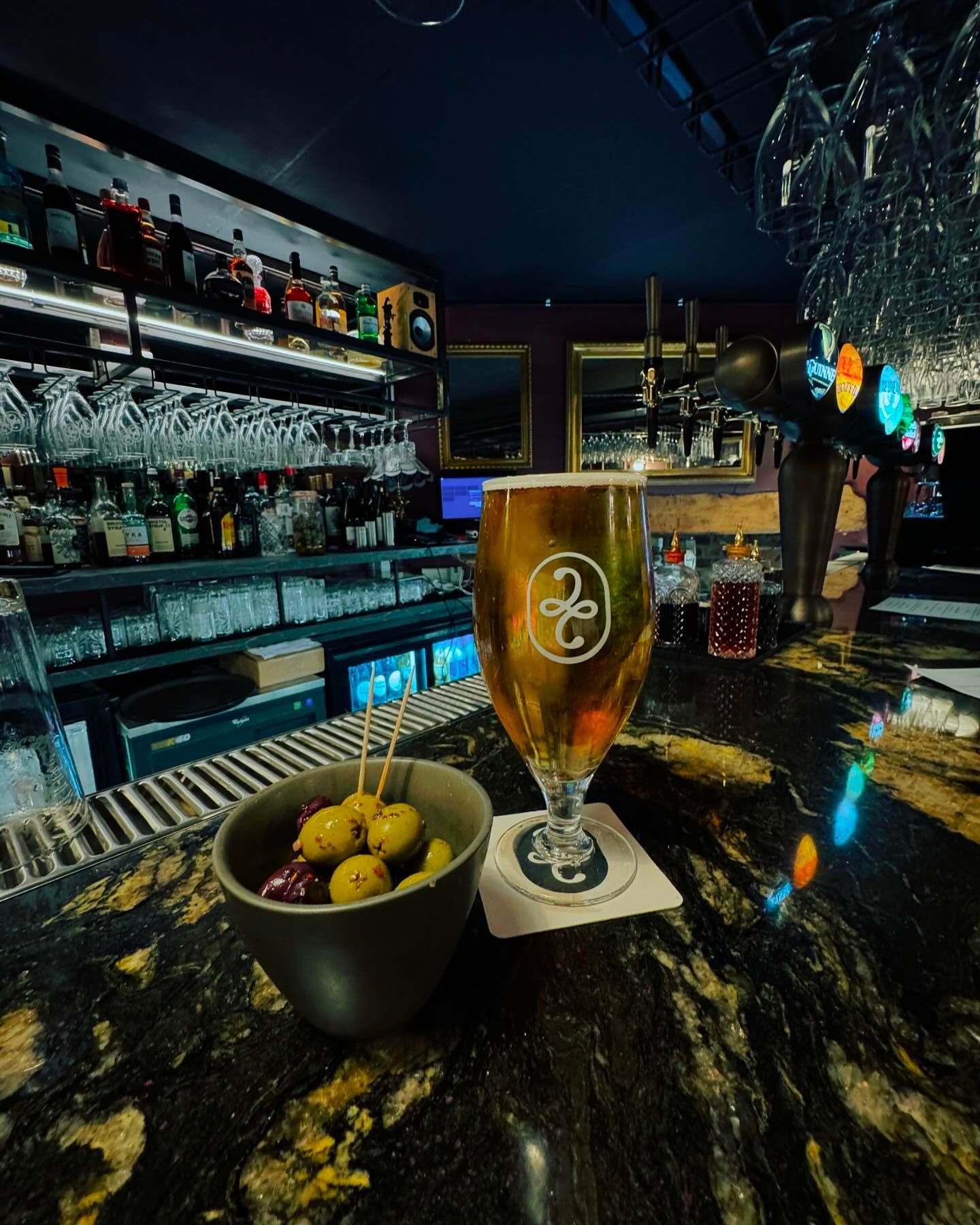 A glass of beer is sitting on a bar next to a bowl of olives.