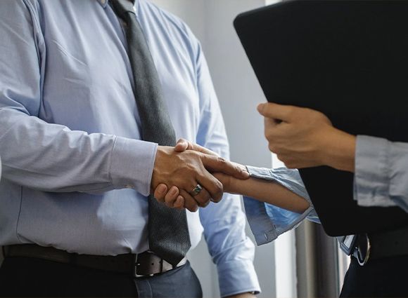 A man and a woman are shaking hands while a woman holds a clipboard.