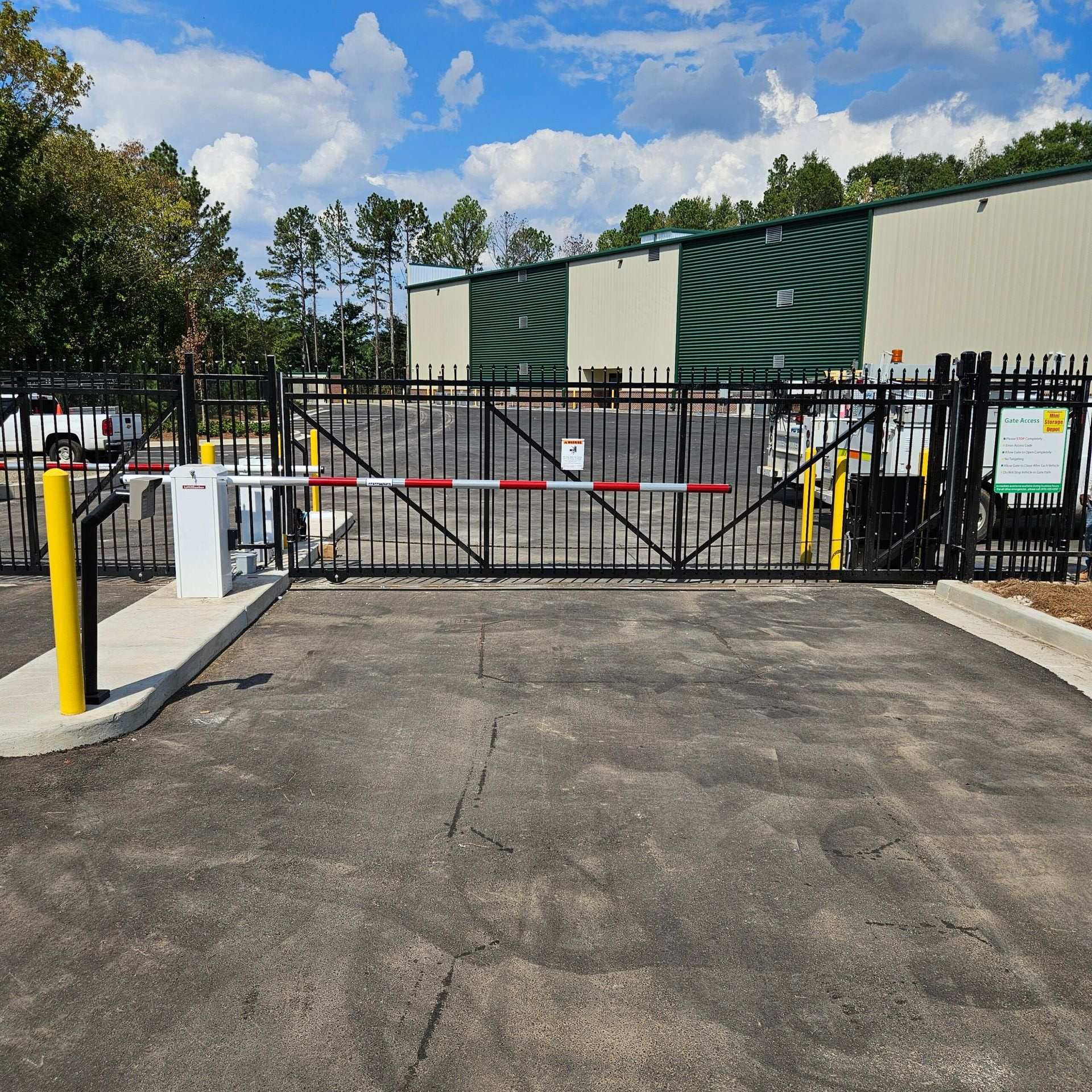 Gated storage facility entrance; black metal gate with red/white arm, gray asphalt, building in background, blue sky.