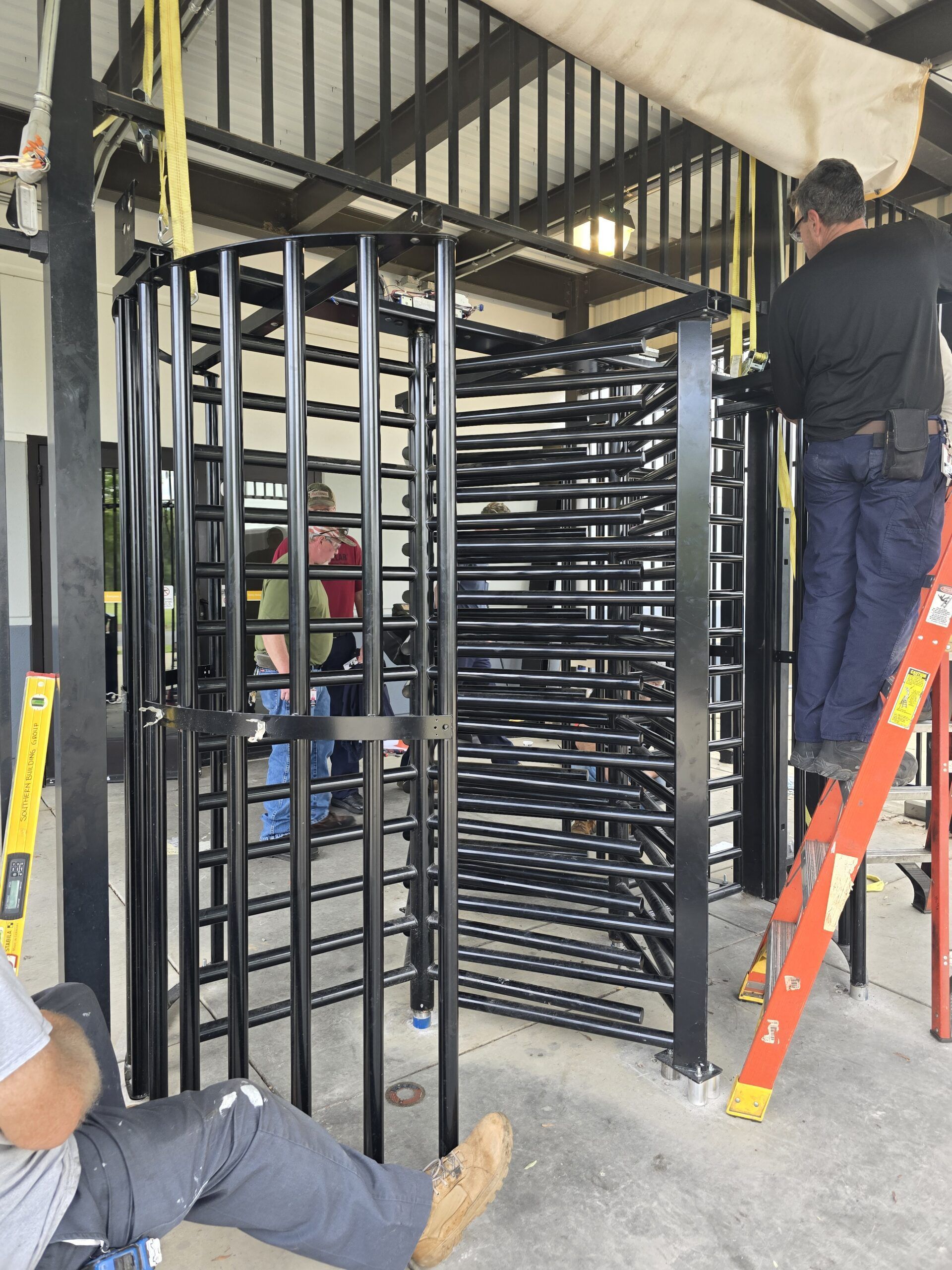 Workers installing a black metal turnstile; one on a ladder, another seated, and a third in the background.