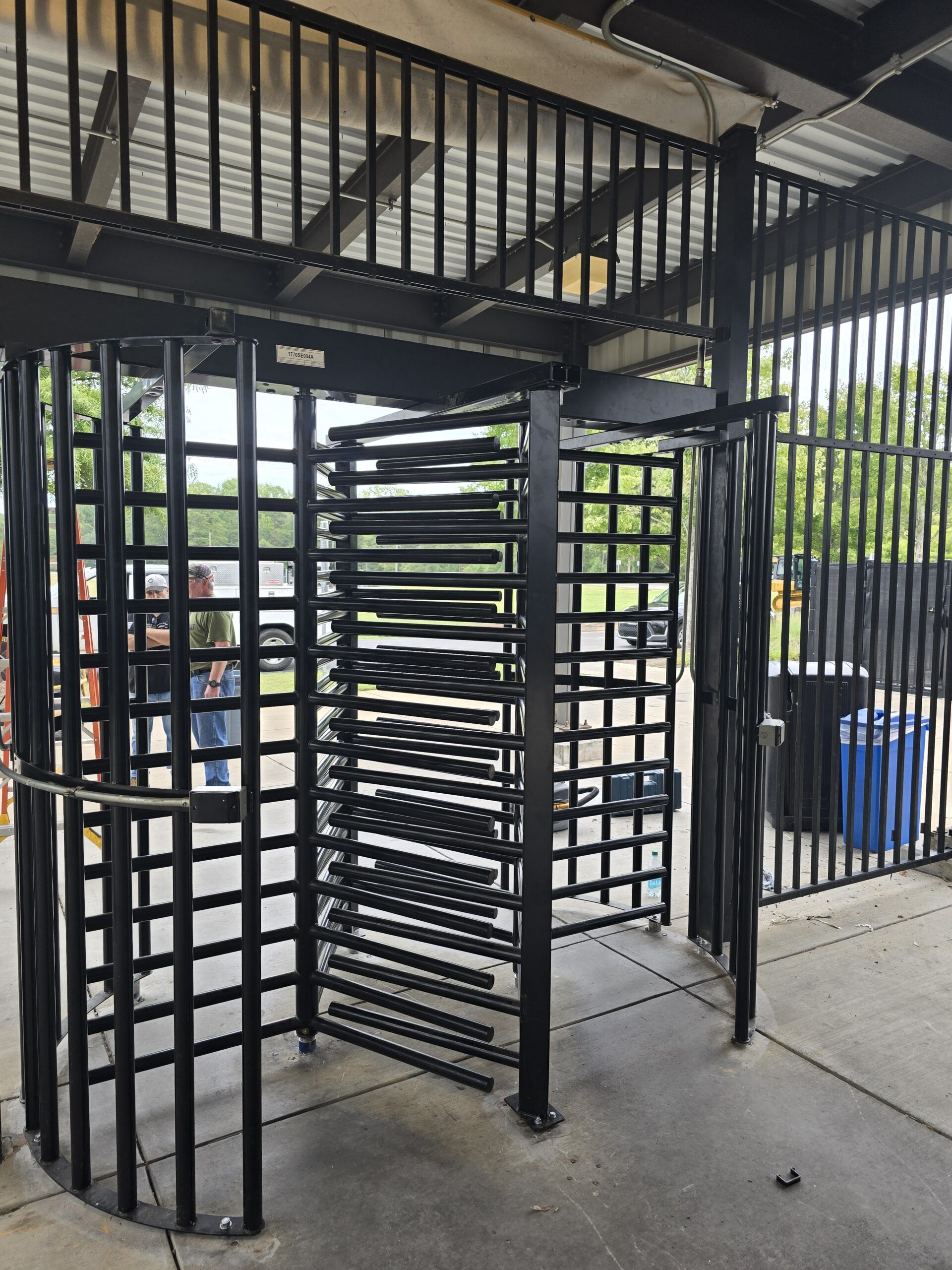 Black turnstile entrance with vertical bars. Metal frame with a corrugated roof in an outdoor setting.