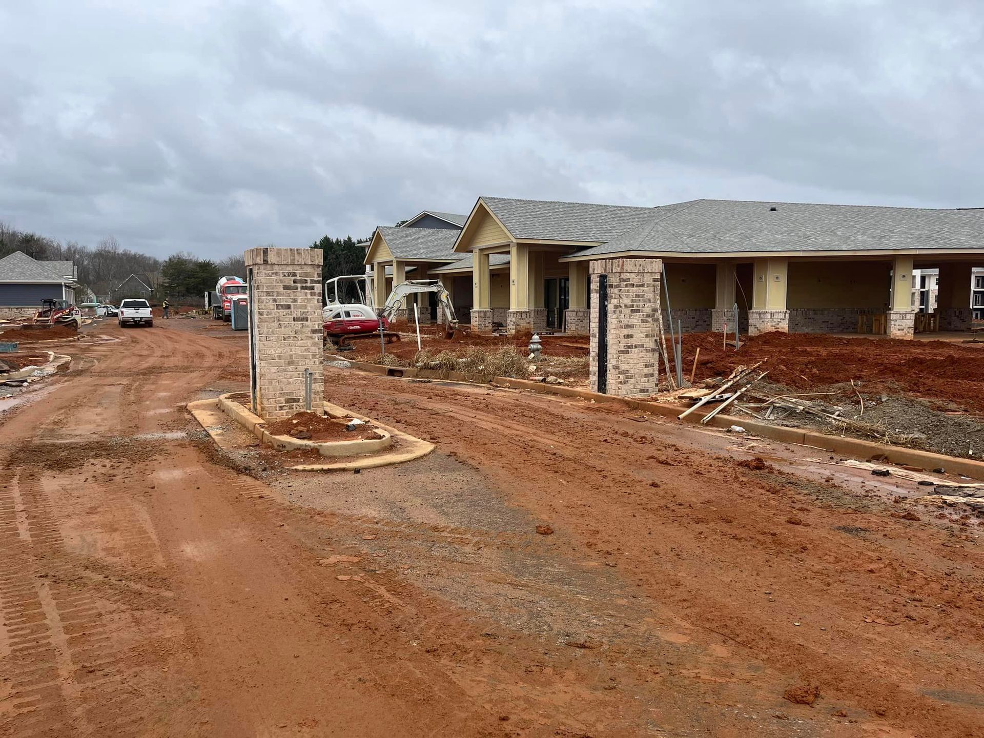 Construction site with red dirt road and partially built buildings. Overcast sky.
