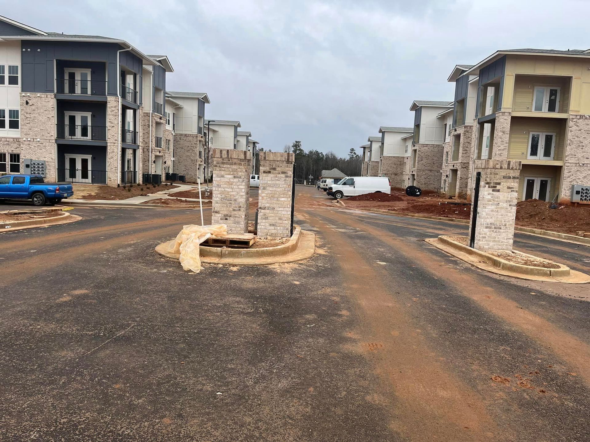 Apartment buildings under construction, entry gate with brick pillars and dirt road.
