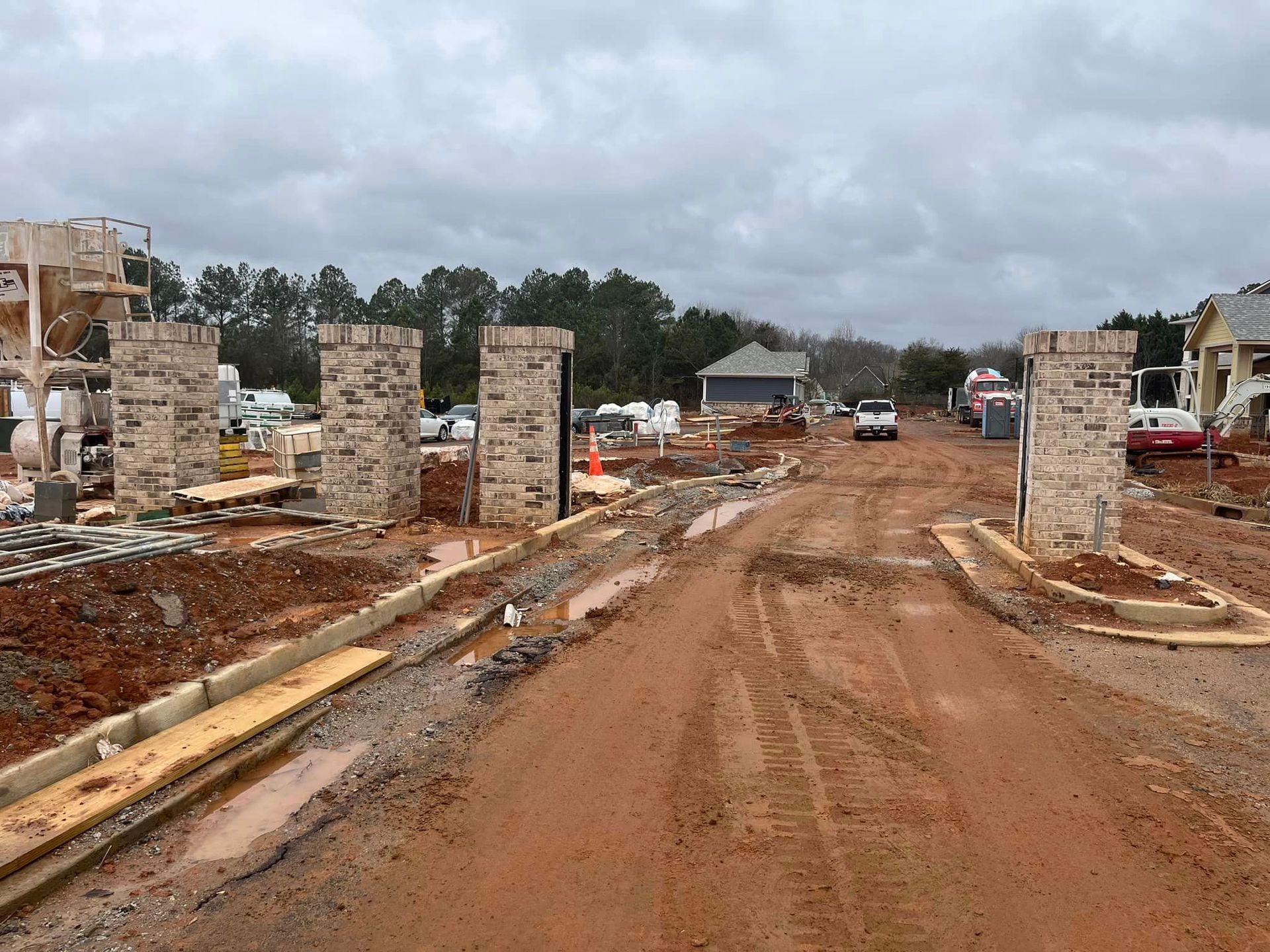 Construction site entrance with brick pillars, dirt road, and houses under construction. Cloudy sky.