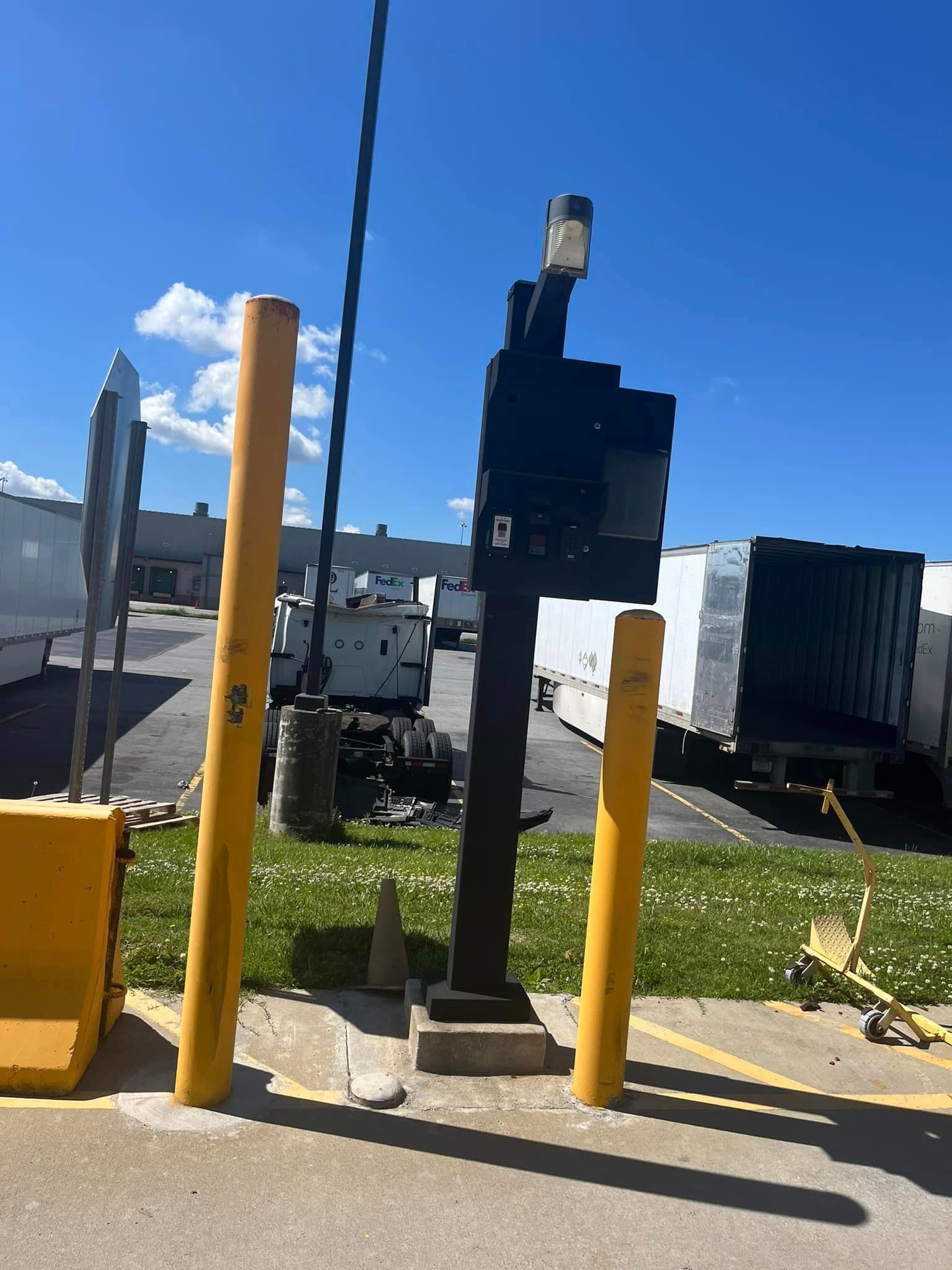 Yellow bollards flank a black industrial post with a light and a camera; trucks in the background.