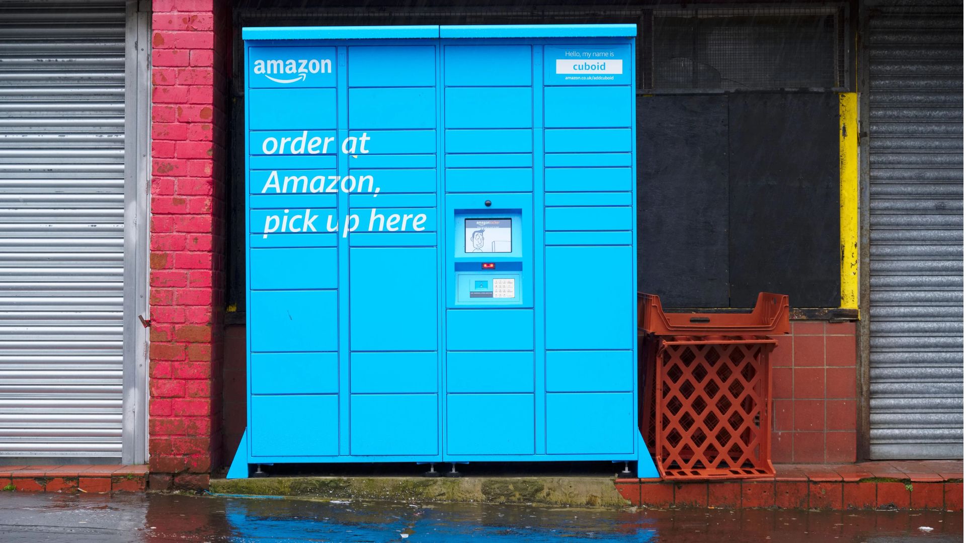 Blue Amazon locker for package pick-up, set in a brick and boarded-up storefront, wet ground.