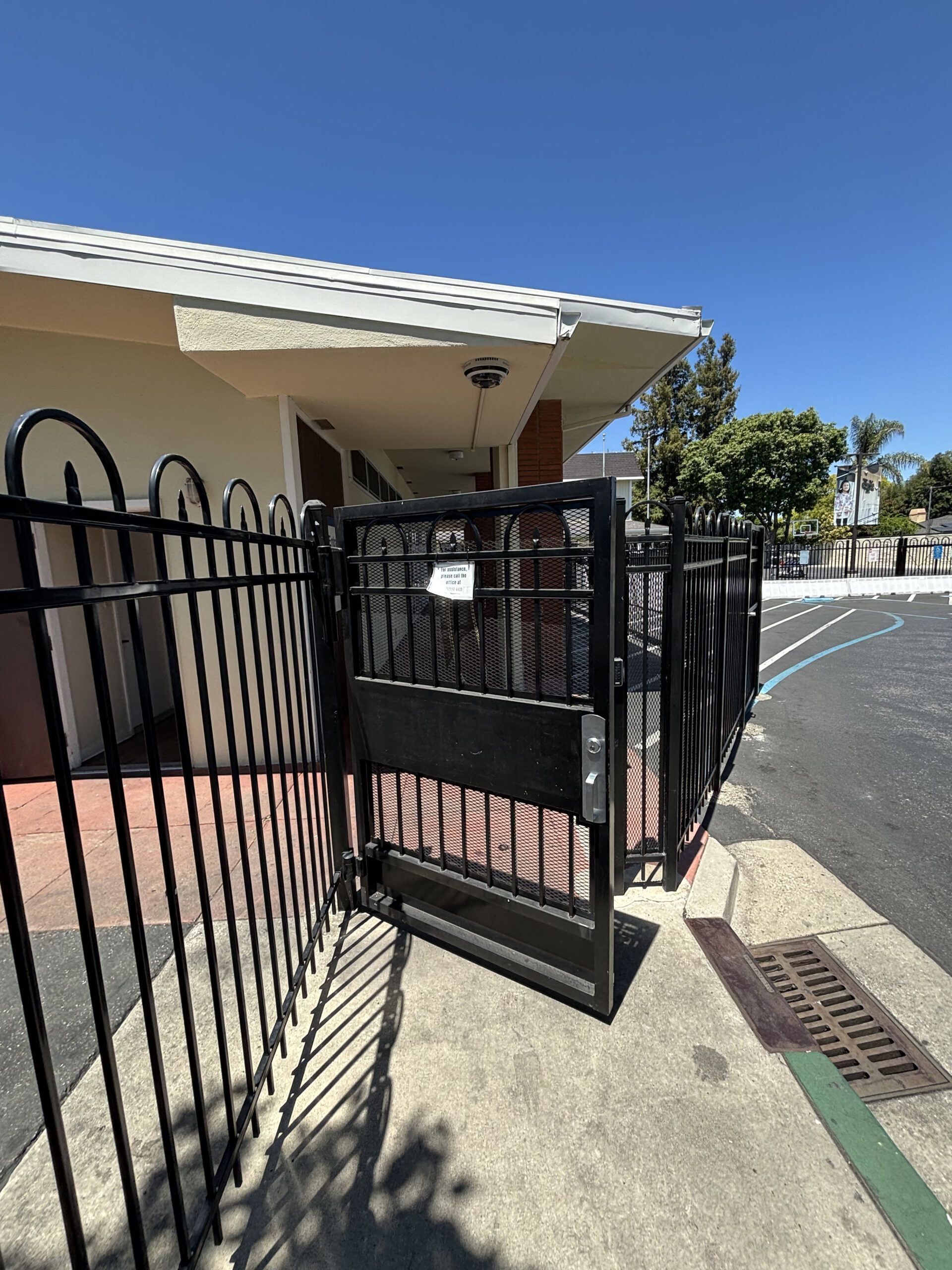 Black metal gate open, leading to building entrance under a white-roofed awning. Sunny day.