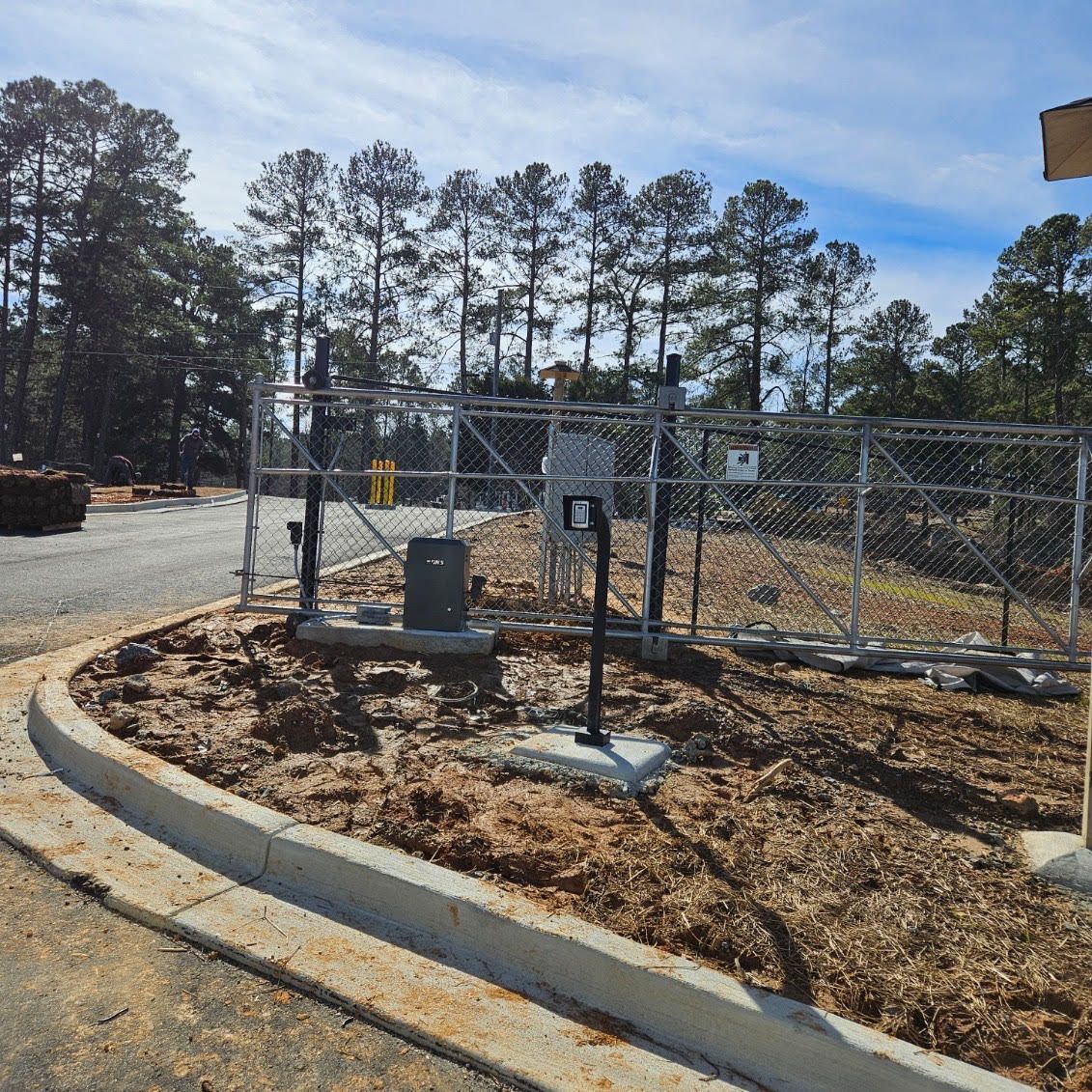 Chain-link fence surrounding an access point, with electronic gate, concrete curbing, and trees in the background.