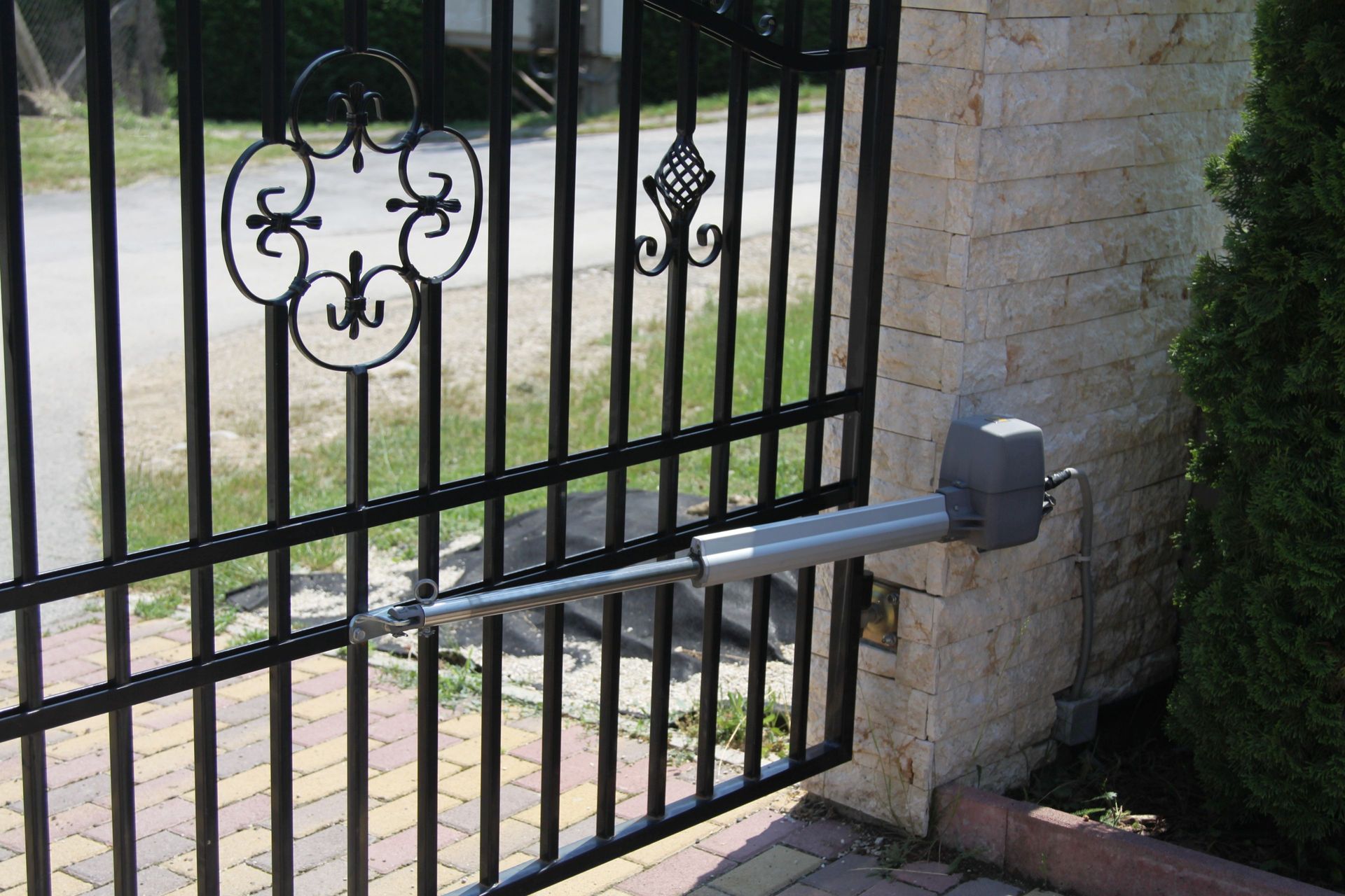 A black wrought-iron gate with an automatic arm opener attached to a light-colored stone pillar.