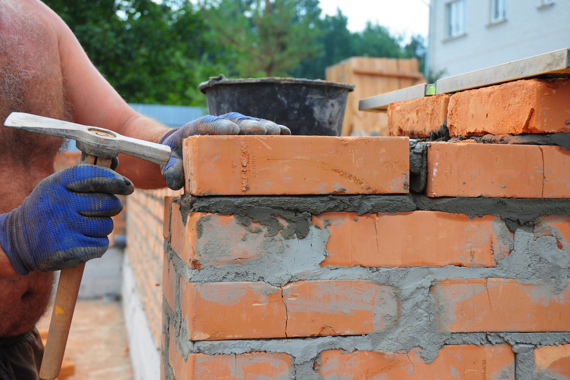 Construction worker laying bricks; mortar between; wearing gloves and holding a hammer.
