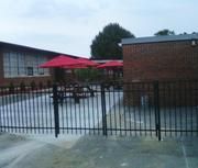 A ornamenntal aluminum and steel fence surrounds a patio area with red umbrellas in front of a brick building.