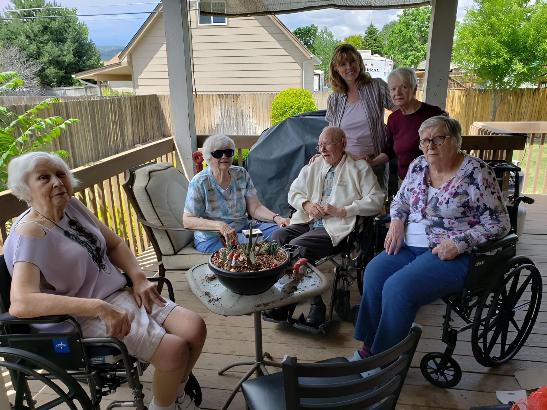 Group of seniors on a wooden deck; some in wheelchairs, smiling. Sunny day with a house and greenery in the background.