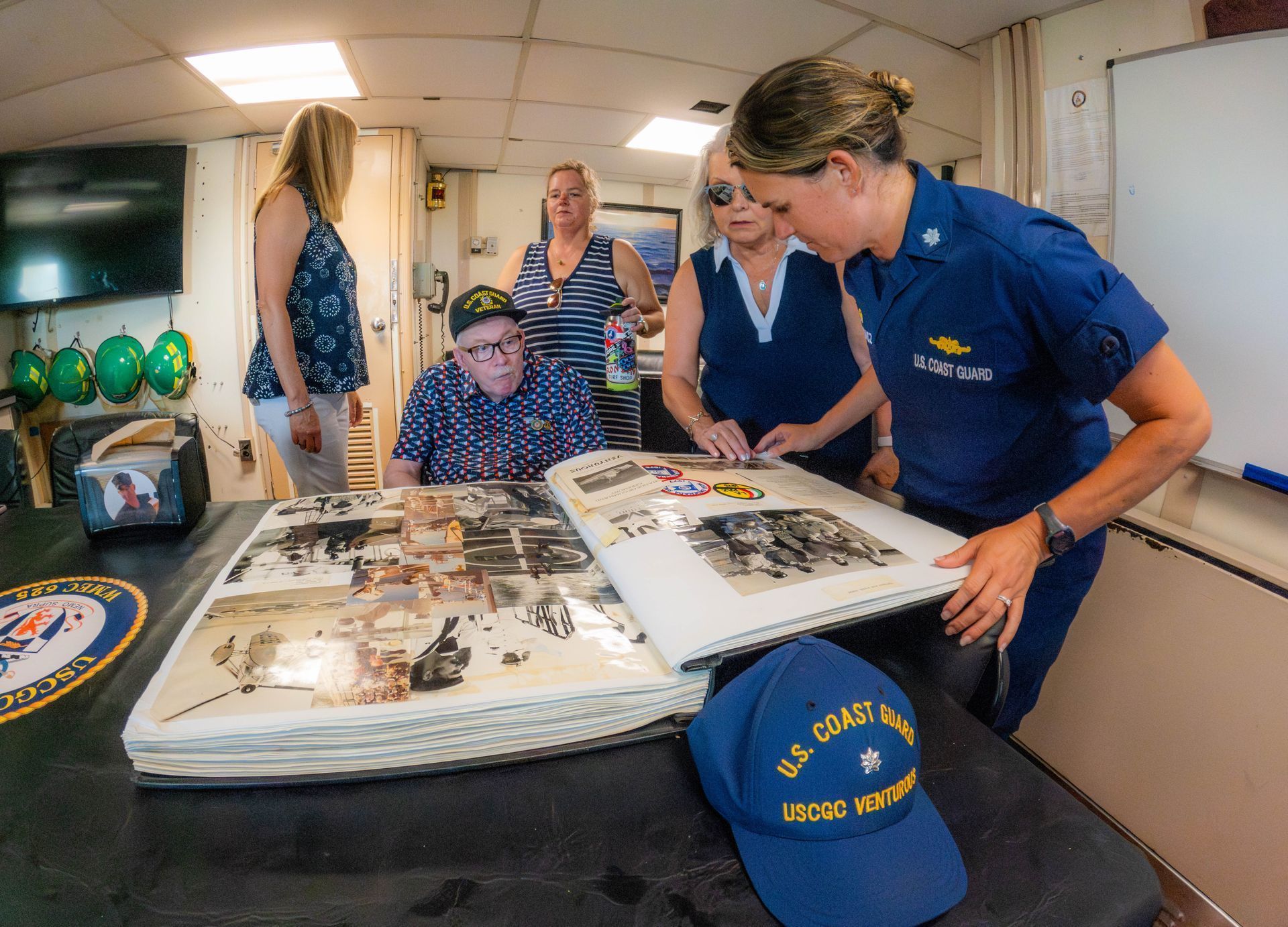 Group of people looking at a scrapbook on a table; a Coast Guard officer points.