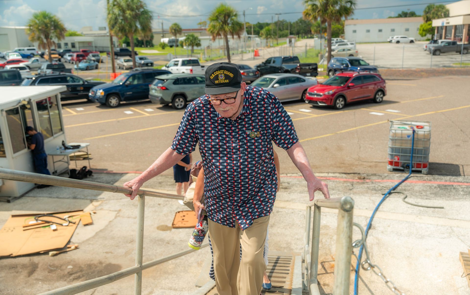 Elderly man in patterned shirt climbs stairs outside, holding handrails. Cars and building in background.