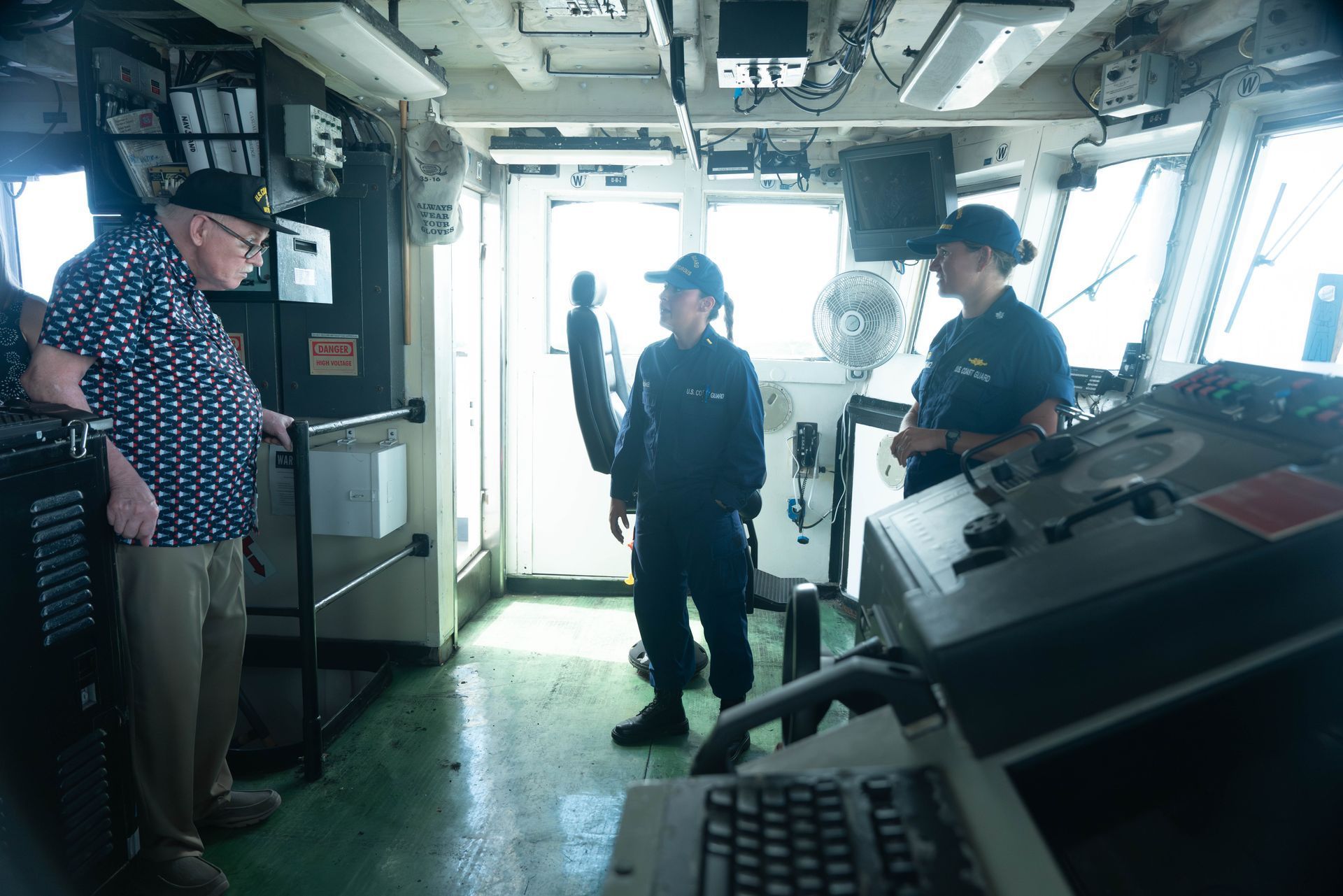 Man in patterned shirt and two uniformed Coast Guard personnel inside a ship's bridge.
