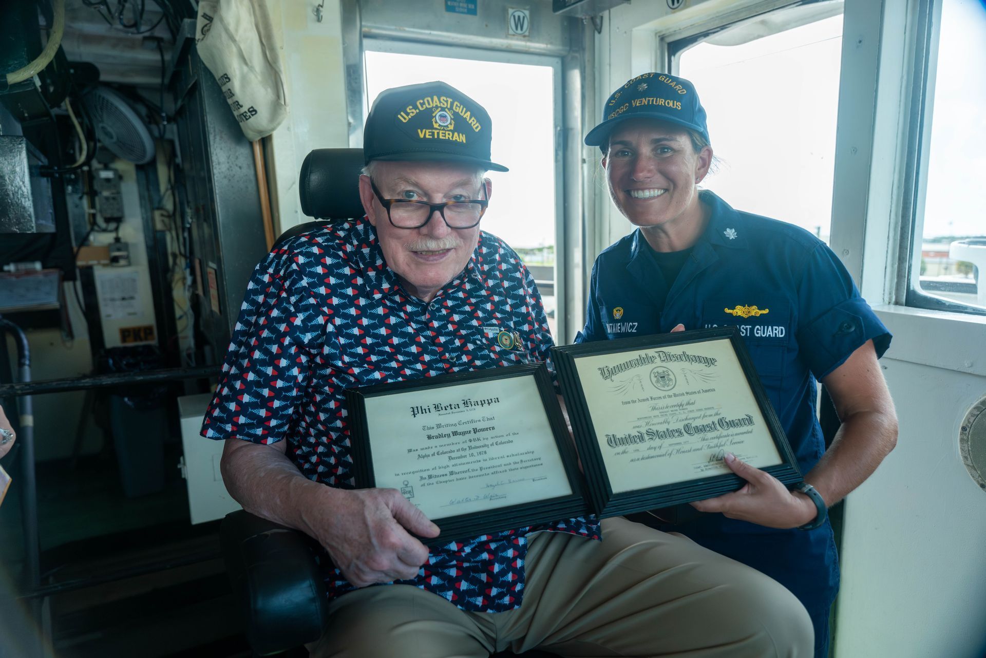 Man and Coast Guard officer, holding framed certificates, inside a ship's bridge.