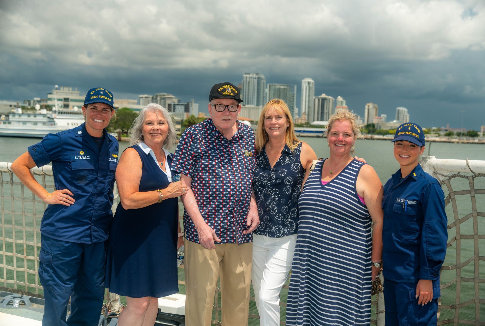 Six people pose on a ship deck with a city skyline in the background; two Coast Guard members and civilians.