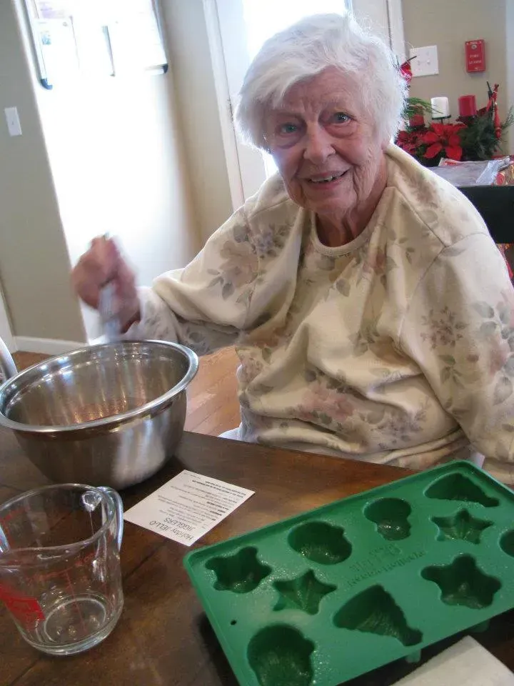 Elderly woman smiling while mixing ingredients, likely baking in a kitchen setting. Cookie mold and measuring cup visible.
