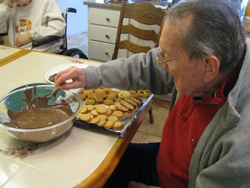 Elderly man dipping cookies in chocolate. Indoor setting, he's wearing glasses and a red shirt.