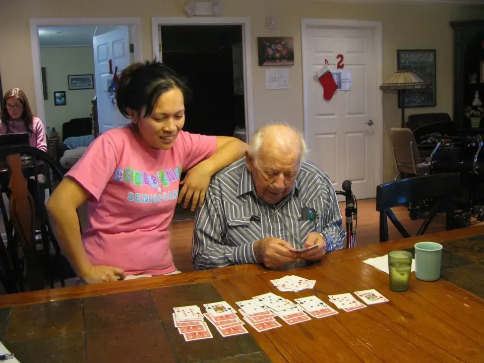 Woman in pink shirt smiles, assisting elderly man playing cards at a table. Home setting.