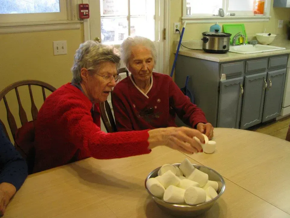 Two older women at a table, building with marshmallows. Kitchen background, one points, both wearing red.