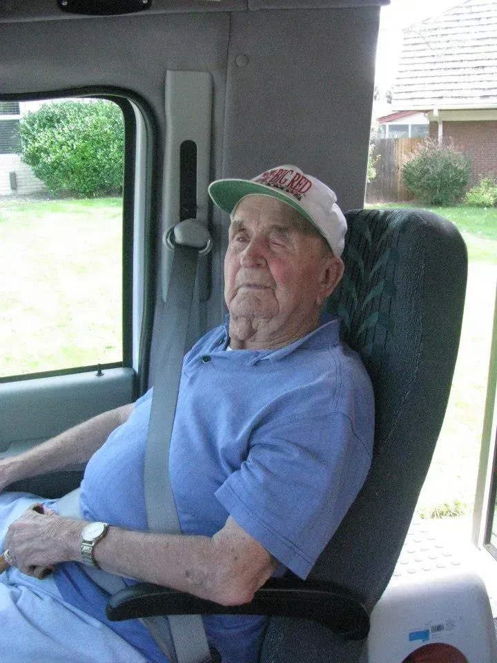 Older man wearing a hat and blue shirt, seated in a vehicle, eyes closed, wearing a seatbelt.