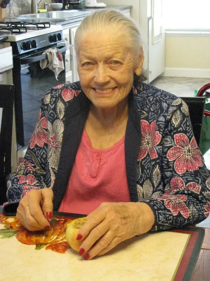 Elderly woman seated at a table, smiling, wearing a pink top and floral blue jacket, holding an apple in a kitchen.