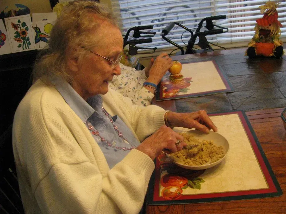 Elderly woman at table eating from a bowl. She wears glasses, a white sweater, and has a focused expression.
