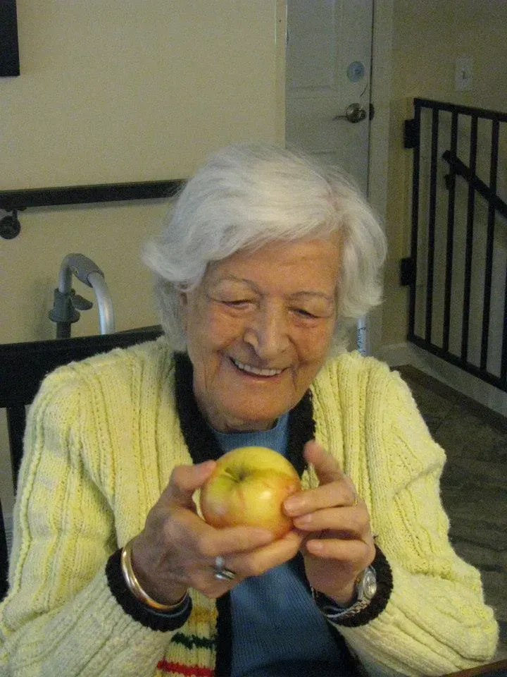 Elderly woman smiles, holding a yellow apple. She wears a yellow sweater, sitting indoors.
