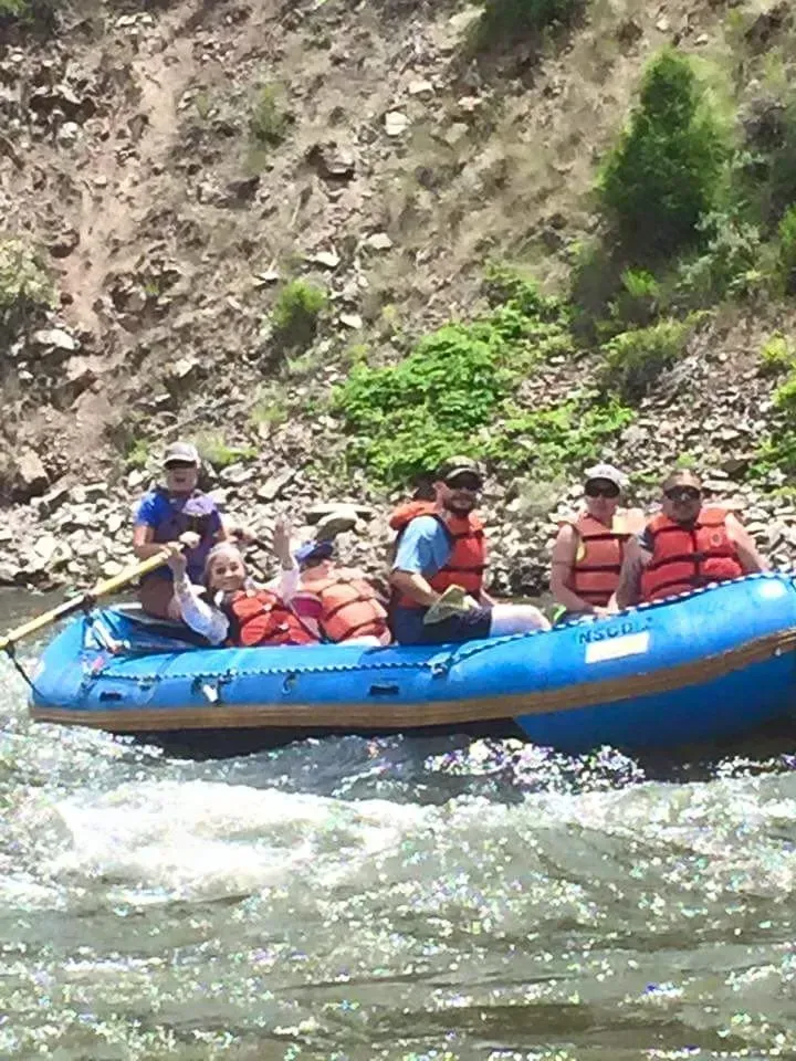 Group of people rafting on a blue raft, waving in a river near a rocky hillside. They are wearing life vests.