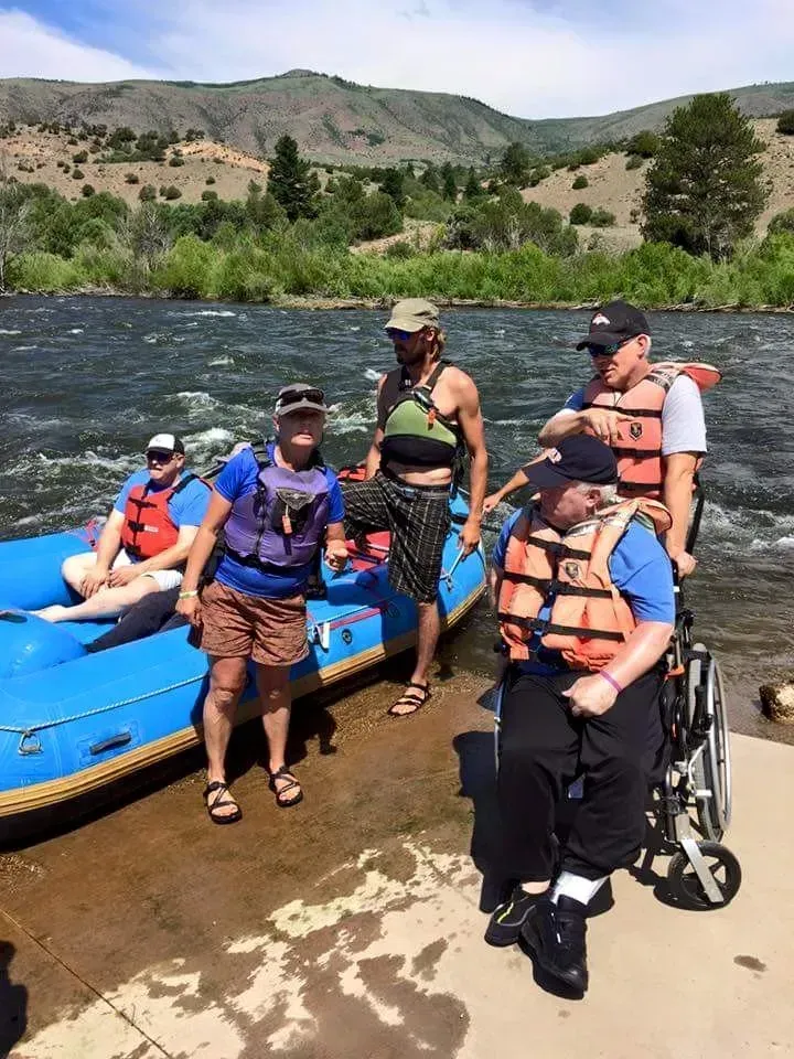 People in life vests near raft, one in wheelchair, on riverbank with mountain backdrop.