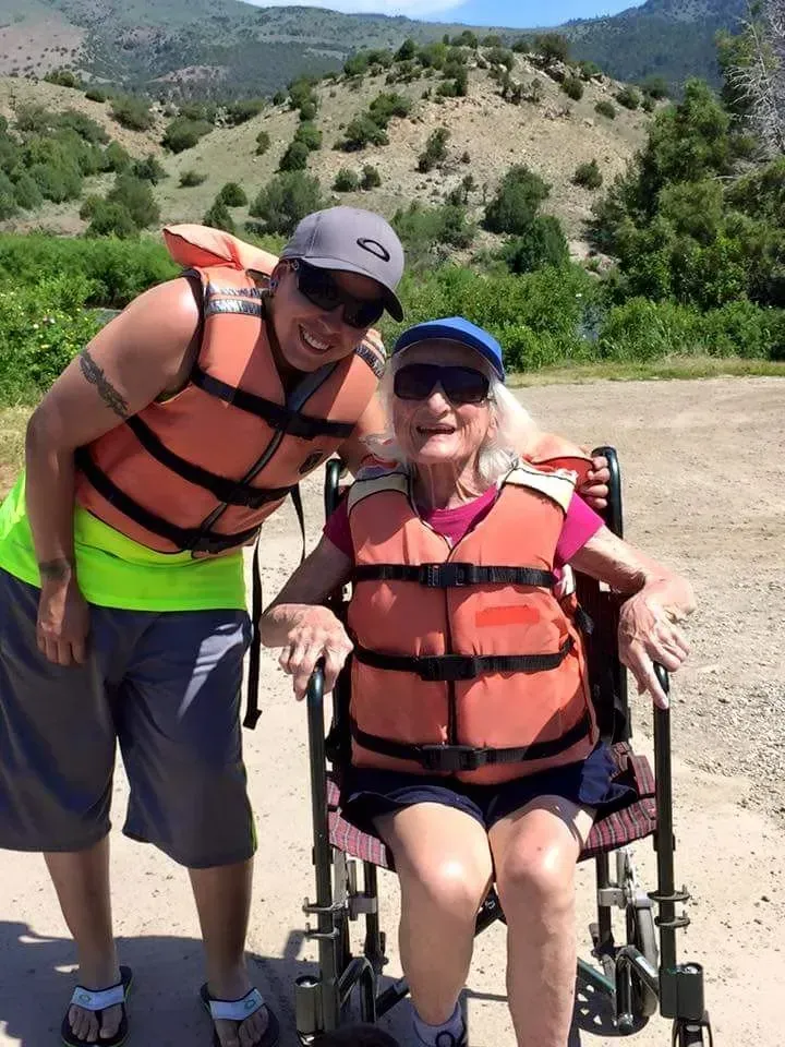 Two people in life vests pose outdoors. A younger person smiles next to an older person in a wheelchair.