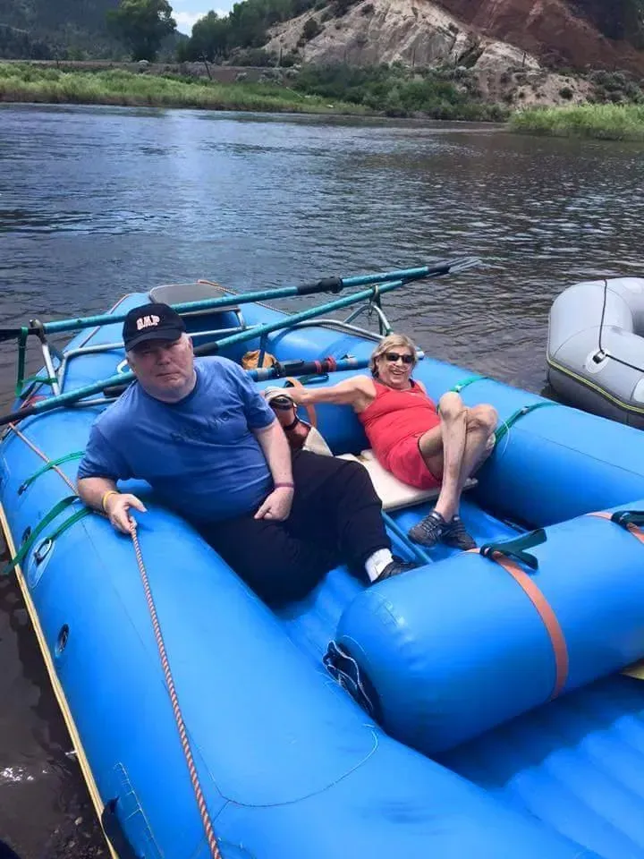 Two people in blue raft on river; man in blue shirt, woman in red dress, mountains in background.