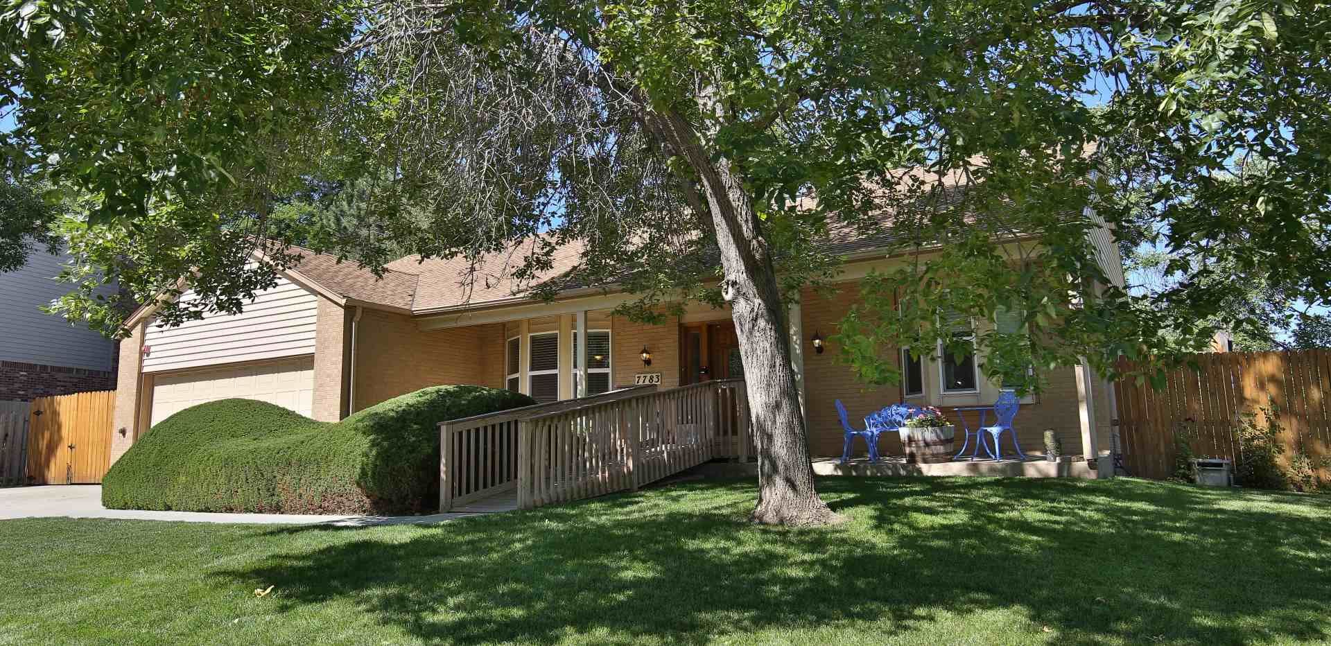 A house with a ramp and a tree. Green grass and blue chairs on a porch.