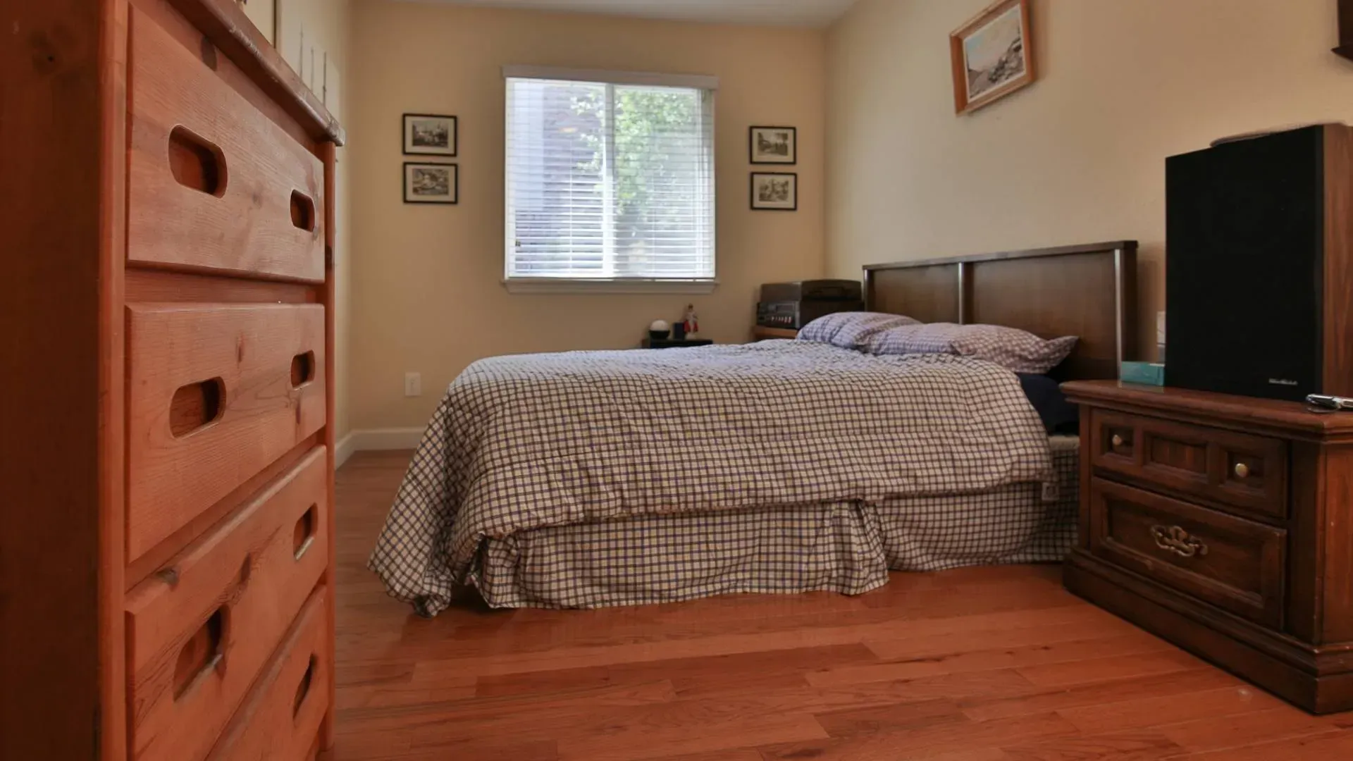 Bedroom with wooden furniture, bed with checkered comforter, window, and artwork on beige walls.