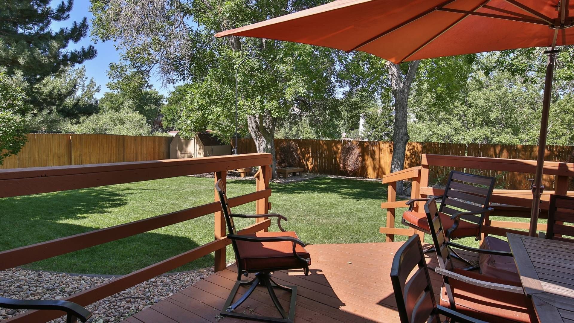 A sunny backyard deck with seating and an orange umbrella, overlooking a fenced yard with trees.