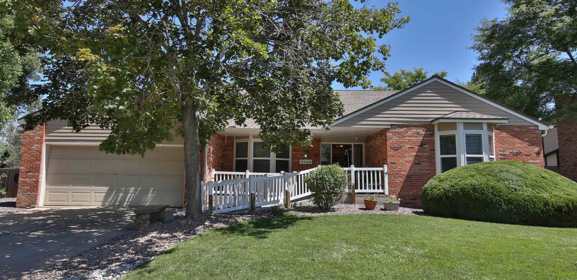 A brick house with a garage and a porch, surrounded by a green lawn and trees on a sunny day.
