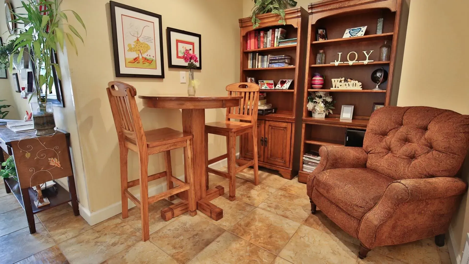 Cozy dining area: wooden table and chairs, bookshelf, and brown recliner chair. Framed art on beige walls.