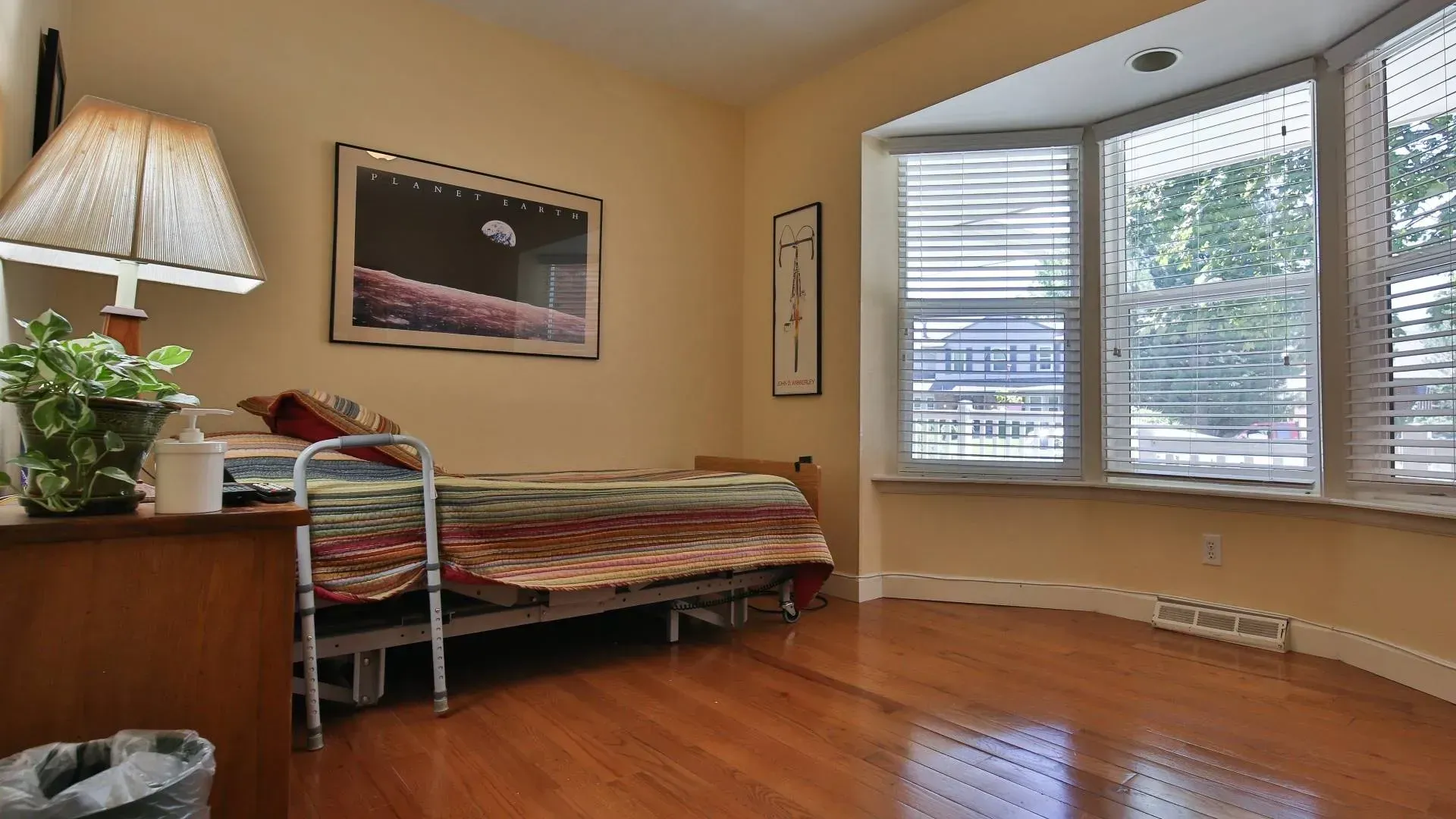 Bedroom with a hospital bed near a bay window with blinds; wood floor.