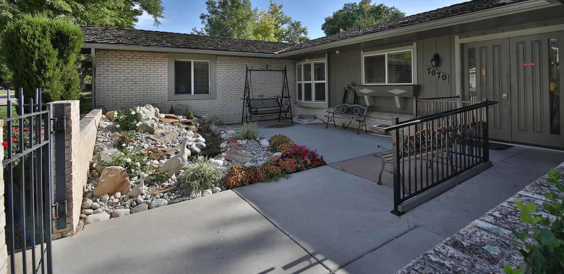 Exterior of a light-colored building with a small garden, two porch swings, and a ramp.