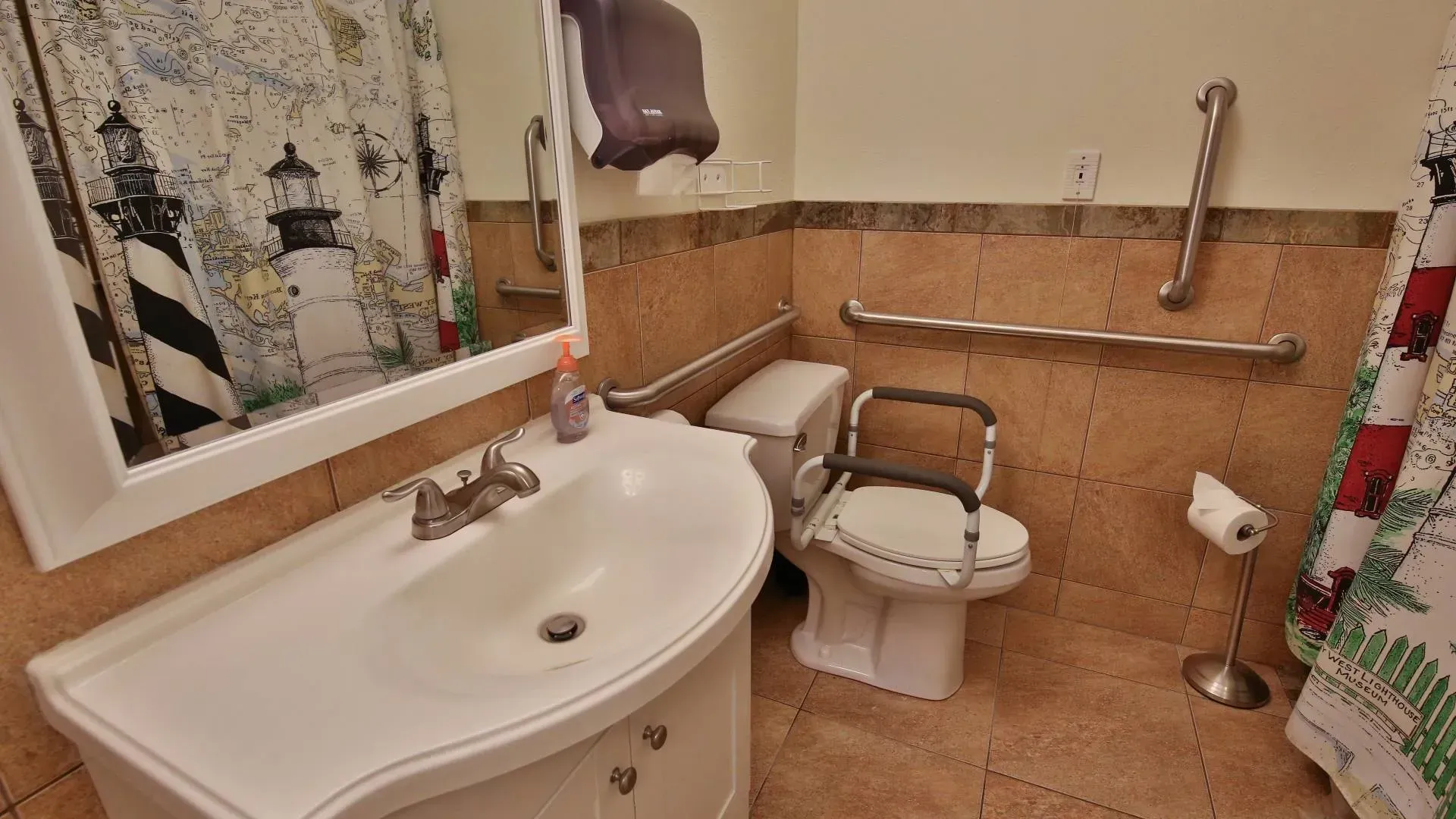 Bathroom with sink, toilet with safety seat, and grab bars. Lighthouse shower curtain and beige tiles.