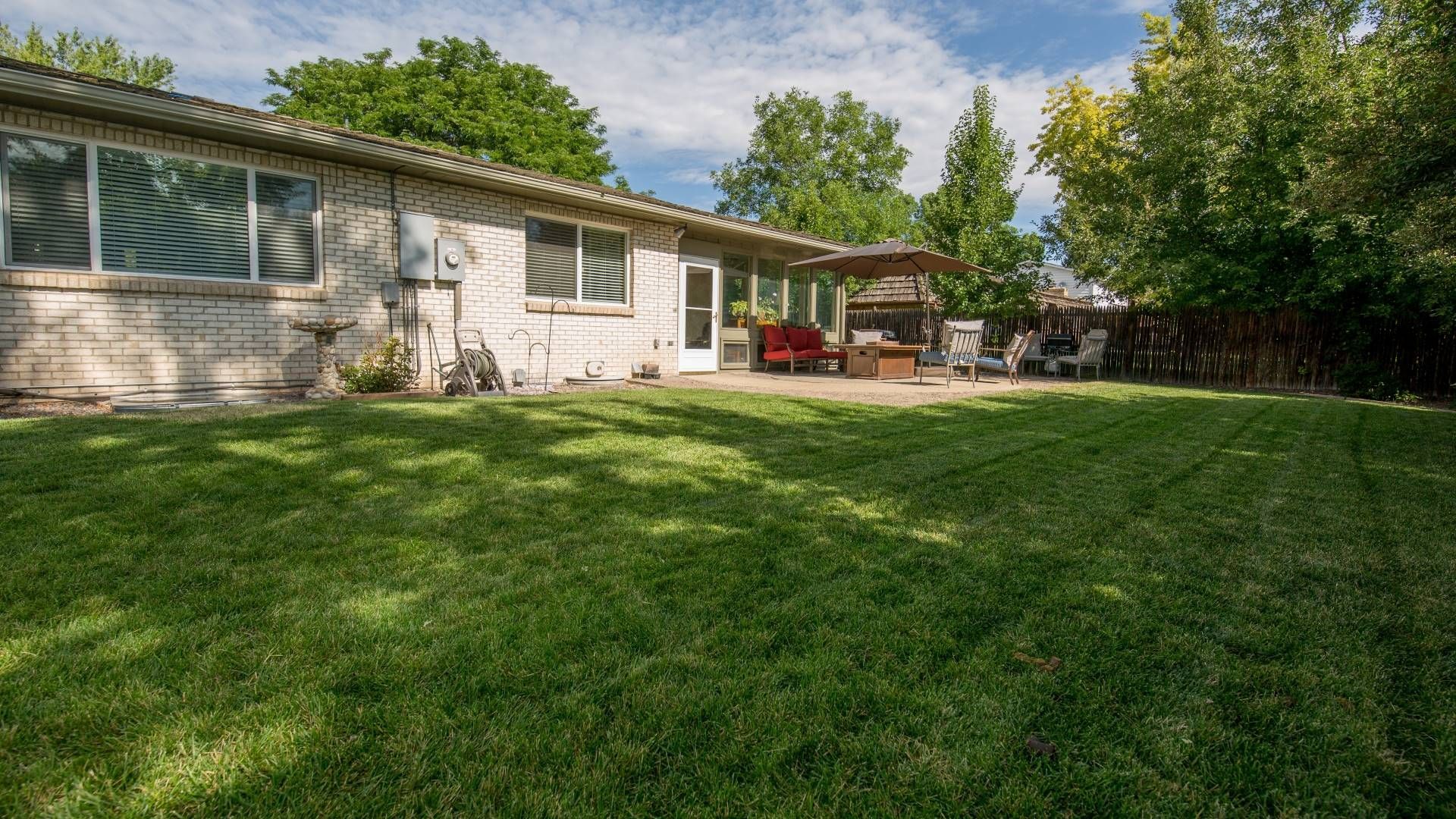 Backyard with green grass, a house, patio with seating, and trees under a blue sky.