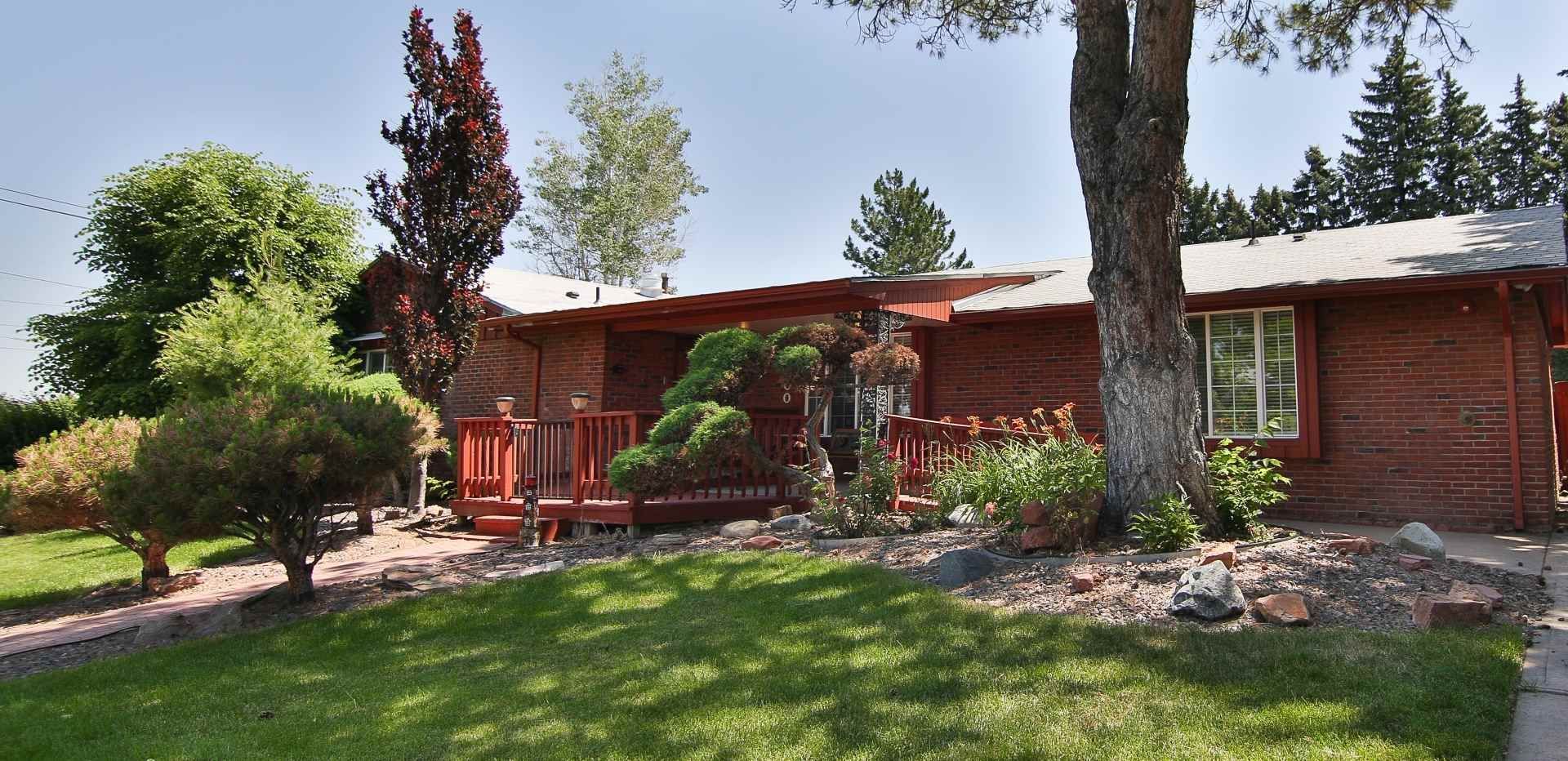 Red-sided house with porch and lush green lawn and trees in front. Sunny day, blue sky.