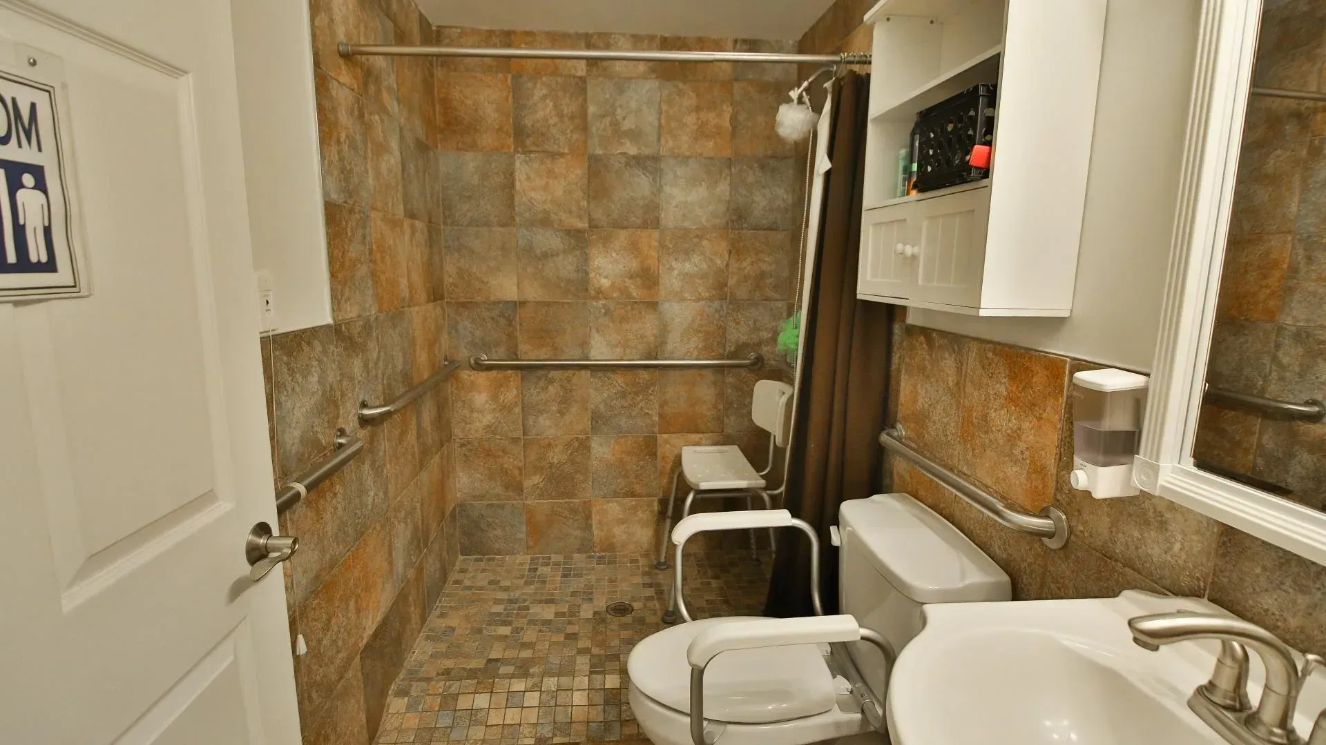 Accessible bathroom with a walk-in shower, grab bars, toilet, and sink, tiled walls in shades of brown and tan.